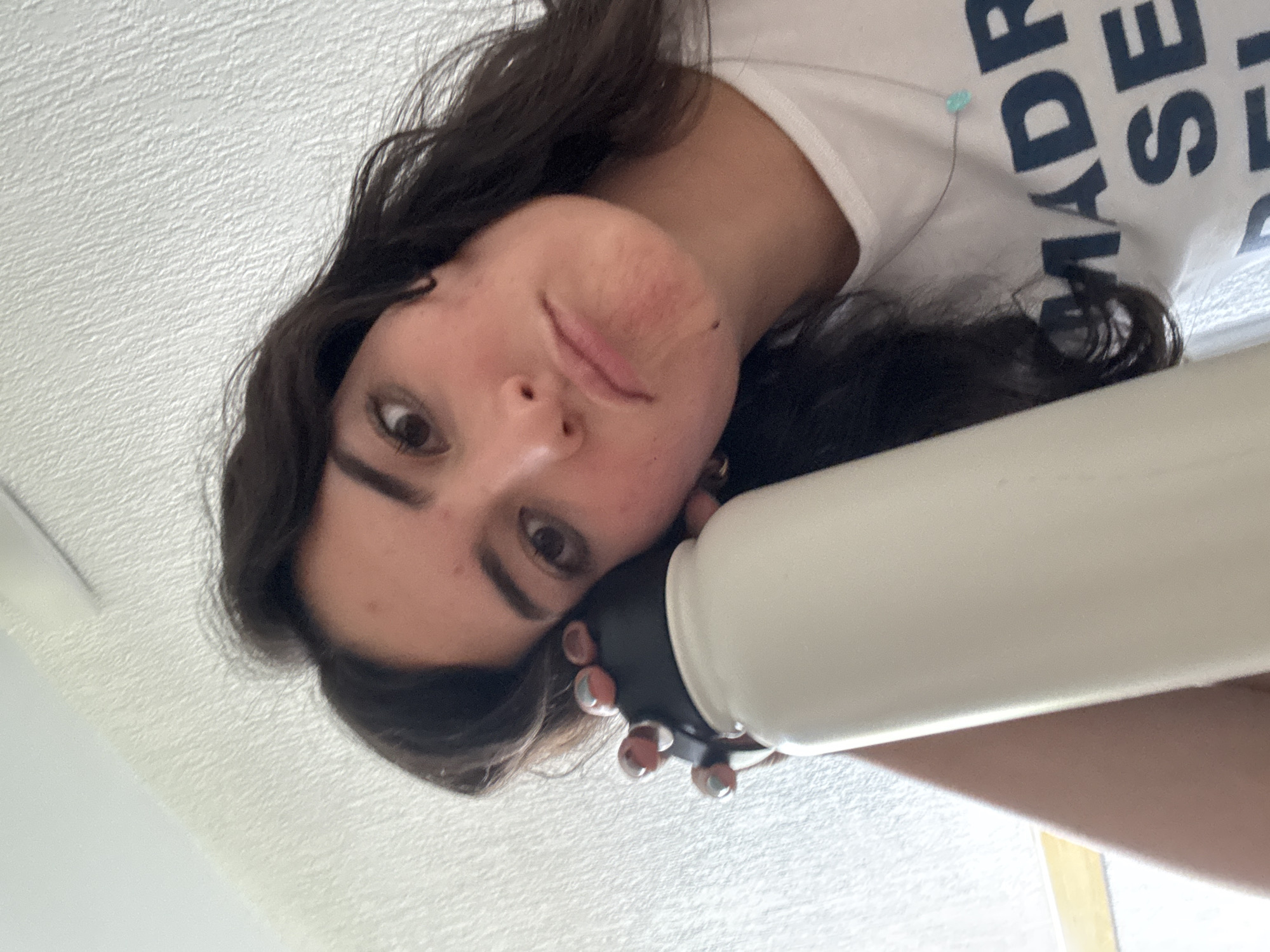 A woman with dark hair and light skin is holding a white water bottle with a black lid up to the camera, obscuring part of her face. She is wearing a white t-shirt with dark letters on it. The background appears to be a white ceiling or wall. The location is San Andrés Cholula, Mexico.