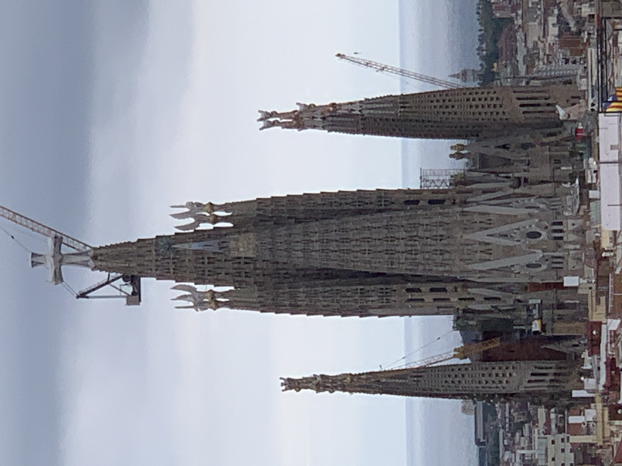 The image shows an aerial view of the Sagrada Familia basilica in Barcelona, Spain. The basilica is a large, unfinished Roman Catholic church, known for its distinctive architecture and intricate detailing. The image captures the basilica's towering spires, some of which are adorned with sculptures and decorative elements. Cranes are visible around the spires, indicating ongoing construction and renovation work. The basilica is surrounded by a cityscape of buildings, and in the background, a hazy horizon suggests the presence of the sea. The sky is overcast with clouds, giving the scene a muted and somewhat dramatic atmosphere.