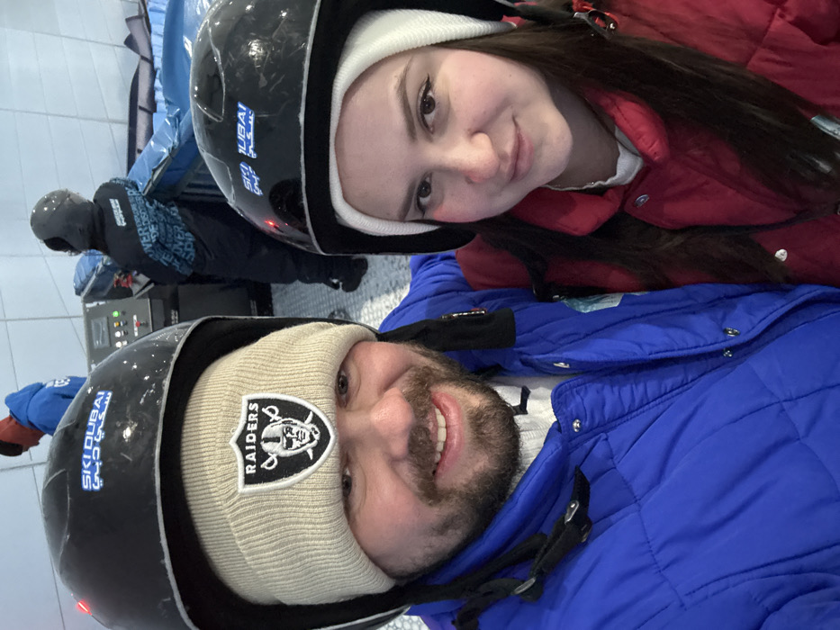 A couple wearing helmets and winter gear poses for a selfie indoors. The woman, on the right, wears a white beanie under her helmet and a red jacket. The man, on the left, wears a Raiders beanie under his helmet and a blue jacket. "Ski Dubai" is visible on the helmets. A third person, wearing a helmet, is seen in the background. The setting appears to be a ski resort or an indoor snow park.