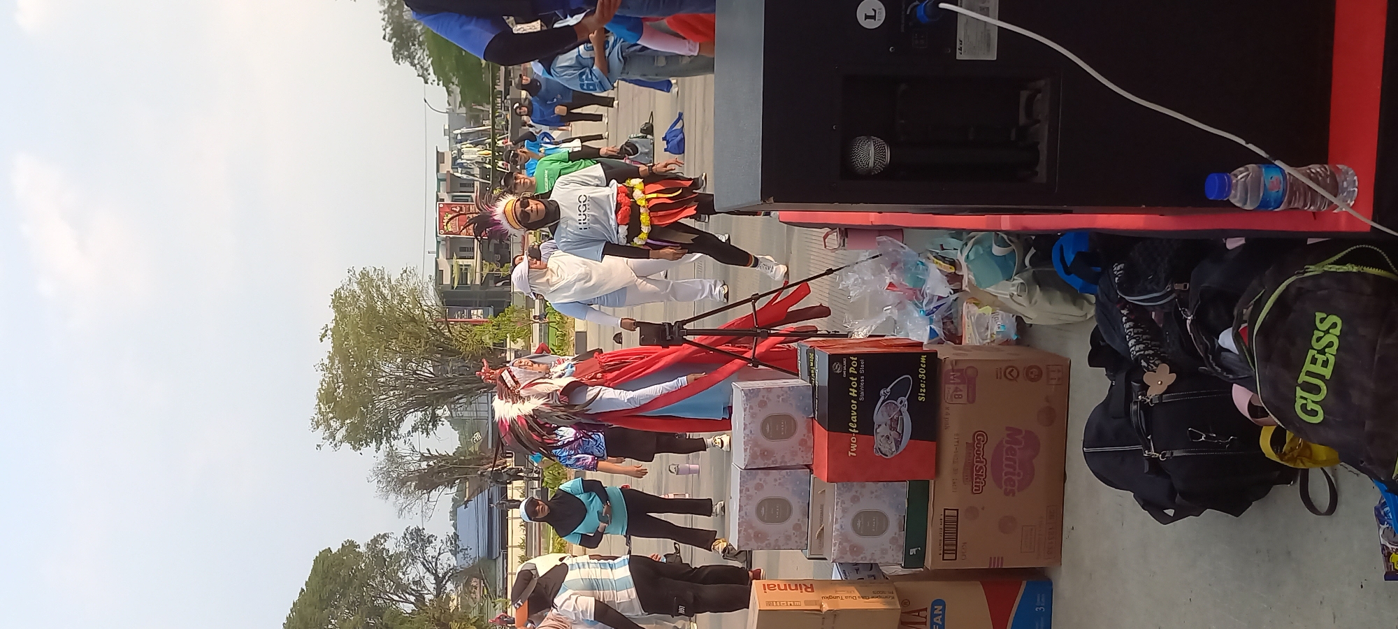 The image depicts an outdoor scene in Melayu, Indonesia, likely a public square or park, with a clear sky and scattered trees in the background. In the foreground, several people are gathered. Some are dressed in athletic wear, suggesting participation in an event like a morning exercise or dance session. One individual stands out, dressed in a costume resembling Native American attire, complete with a headdress. Another person wears a t-shirt with "HUGO" printed on it.
To the right of the frame, a large black speaker is visible, with a microphone plugged into it and a water bottle placed nearby. In the center, a stack of cardboard boxes is present, some containing food items like "Two-flavor Hot Pot." Behind these boxes, a tripod holds a smartphone, possibly for recording the event.
A collection of bags, including backpacks and duffel bags, are scattered on the ground to the left of the speaker, suggesting attendees have brought their belongings. The overall atmosphere appears to be casual and communal, with people engaged in an activity outdoors. The lighting suggests it is daytime, likely morning or late afternoon.