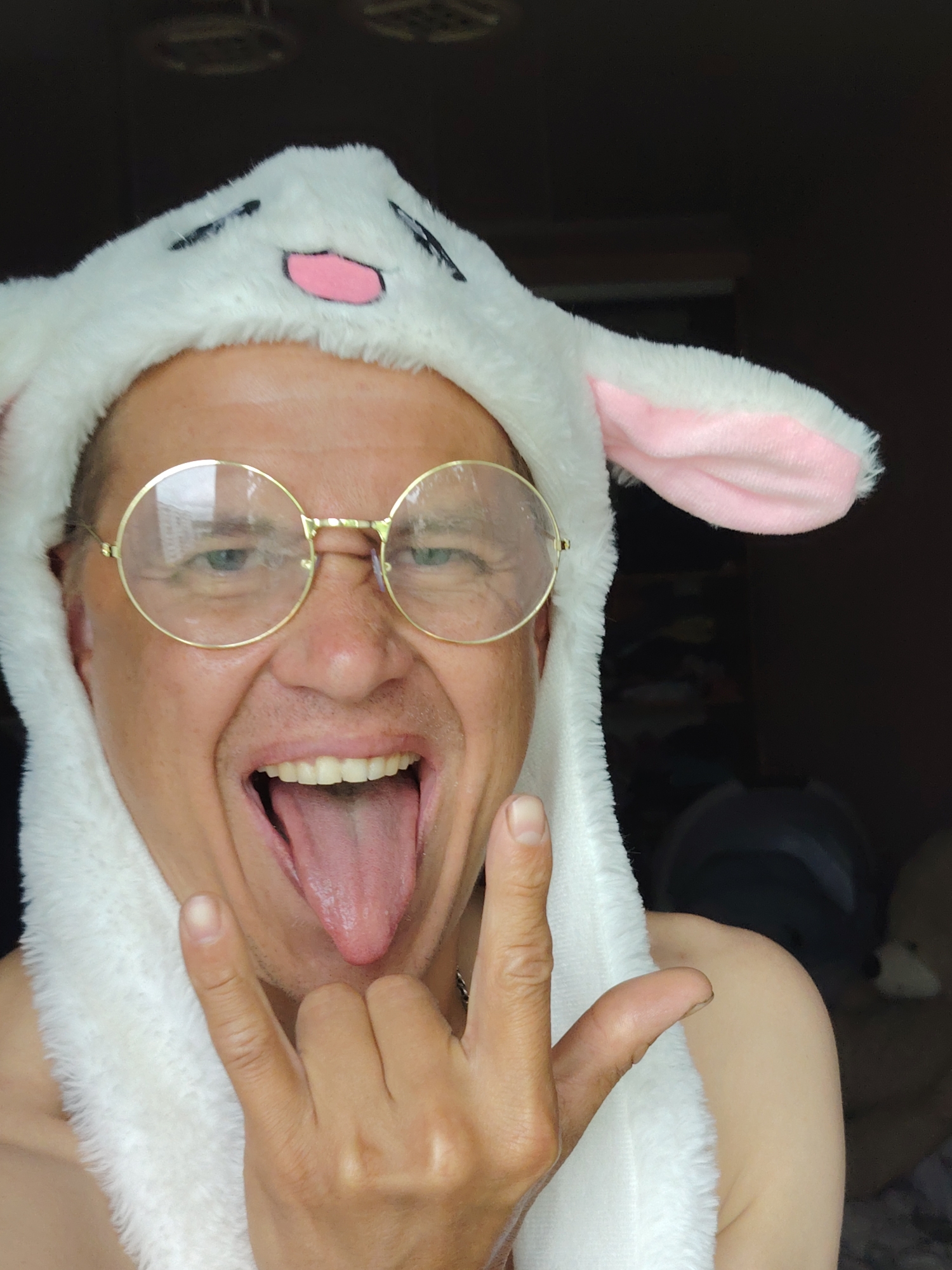 A close-up, selfie-style portrait shows a light-skinned person wearing a white plush bunny hat with pink ears and facial features. The person is wearing round, gold-rimmed glasses and has their mouth open with their tongue sticking out. The person is also making a rock-and-roll hand gesture with their right hand, with their index finger and pinky finger extended. The background is dark and appears to be indoors, with some indistinct objects visible. No specific location in Chaikovsky, Russia is identifiable from the image.