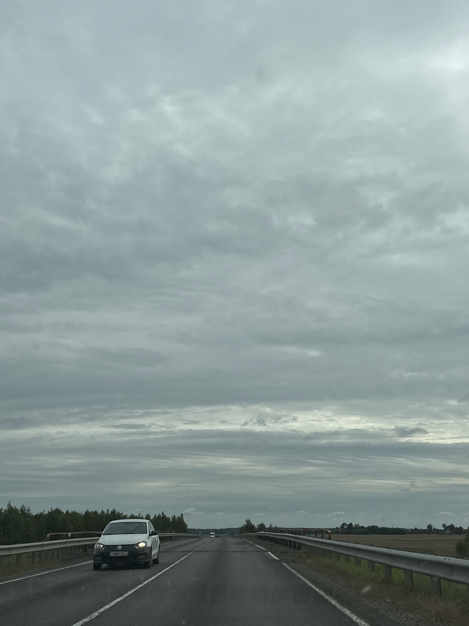The image shows a highway scene in the vicinity of Гарбацэвіцкі сельскі Савет, Belarus, under a cloudy sky. The road is asphalt with white lane markings. A white Volkswagen Polo is traveling in the left lane, heading towards the viewer, with its headlights on. The car has a license plate that reads "9083 EE-6". On the right side of the road, there is a metal guardrail, and beyond that, a field. On the left side of the road, there is a dense line of trees. In the distance, other vehicles and signs of human activity can be observed on the road. The overall atmosphere is overcast and somewhat somber.