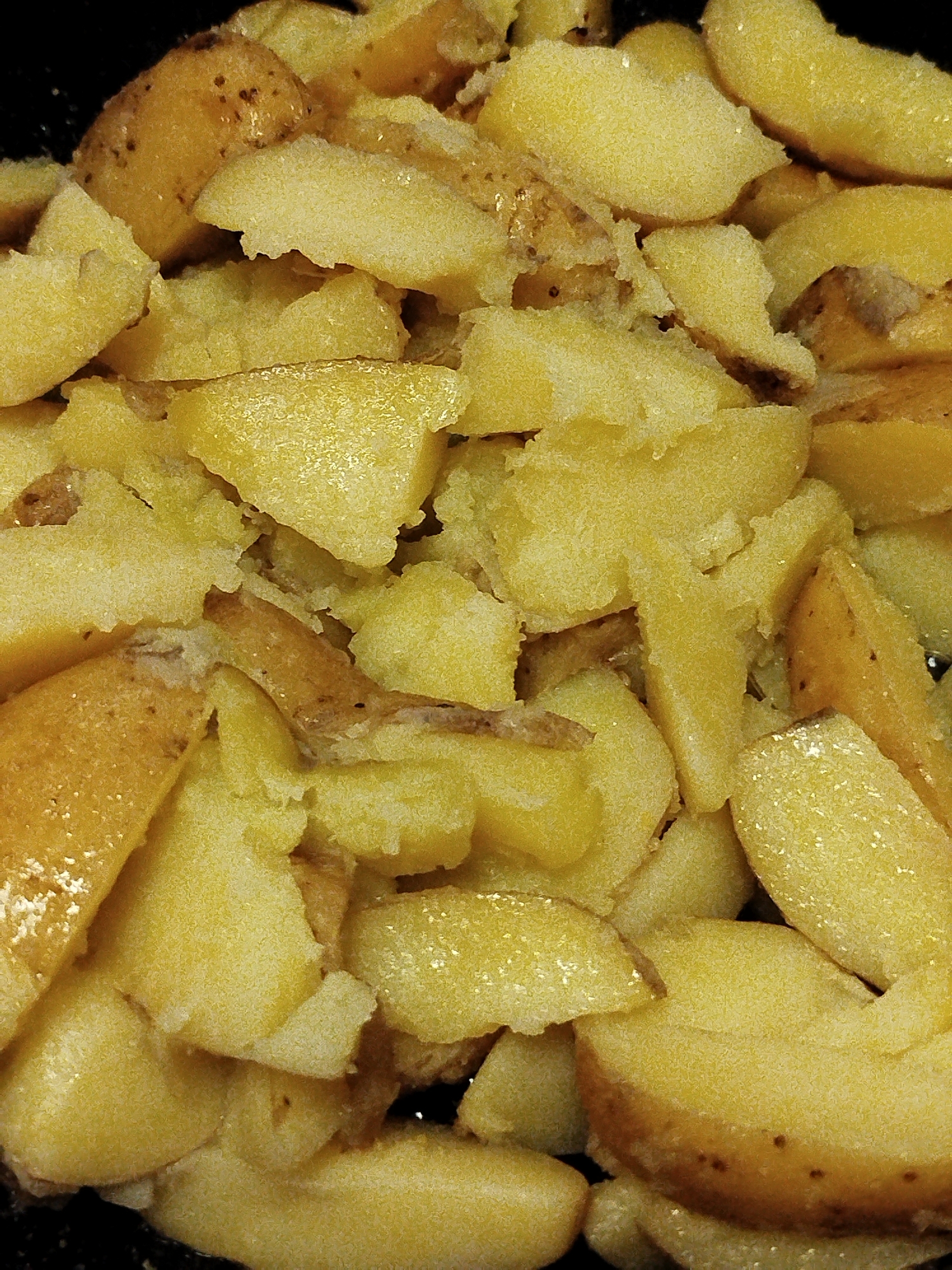 This is a close-up, overhead shot of a pile of golden brown, pan-fried potato wedges. The potatoes are unevenly cut and appear to be seasoned with salt and possibly some oil, giving them a slightly glistening texture. Some potato skins are still attached, adding to the rustic appearance. The lighting is somewhat harsh, creating bright highlights and deep shadows. The background is dark and out of focus, suggesting the potatoes are in a pan or serving dish. There are no people visible in the image, and no indication of the location within Nova Znamianka, Ukraine. The scene depicts a simple, cooked food item.