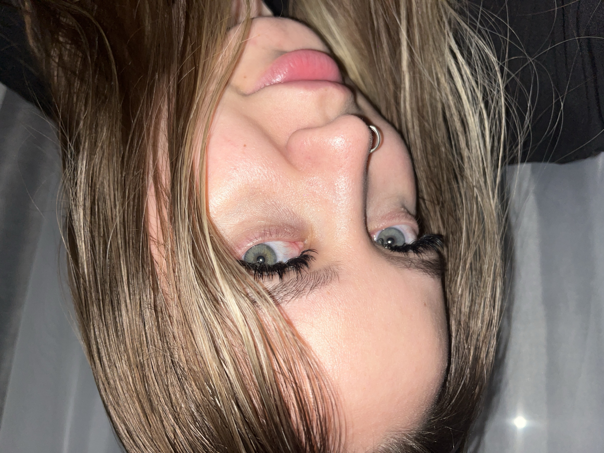 This close-up portrait features an individual, likely a young woman, captured with the image rotated 90 degrees clockwise, presenting the face sideways. Her head appears tilted back, with her gaze directed somewhat upwards.
She has light skin with a smooth complexion. Her eyes are a striking light color, possibly blue or green, with notably long, dark eyelashes, which appear enhanced. One eye is partially obscured by strands of her hair. Her nose is prominent and features a silver hoop nose ring in the right nostril. Her lips are full, naturally pink, and slightly parted.
Her hair is long and straight to slightly wavy, a mix of light brown and distinct blonde highlights, appearing lustrous. It frames her face, with some strands falling across her forehead and eyes.
The background is dark and out of focus, suggesting an indoor setting with artificial lighting. A dark, possibly fabric, element is visible behind her head. The expression is neutral, conveying a direct and steady gaze. There is no visible text in the image.
