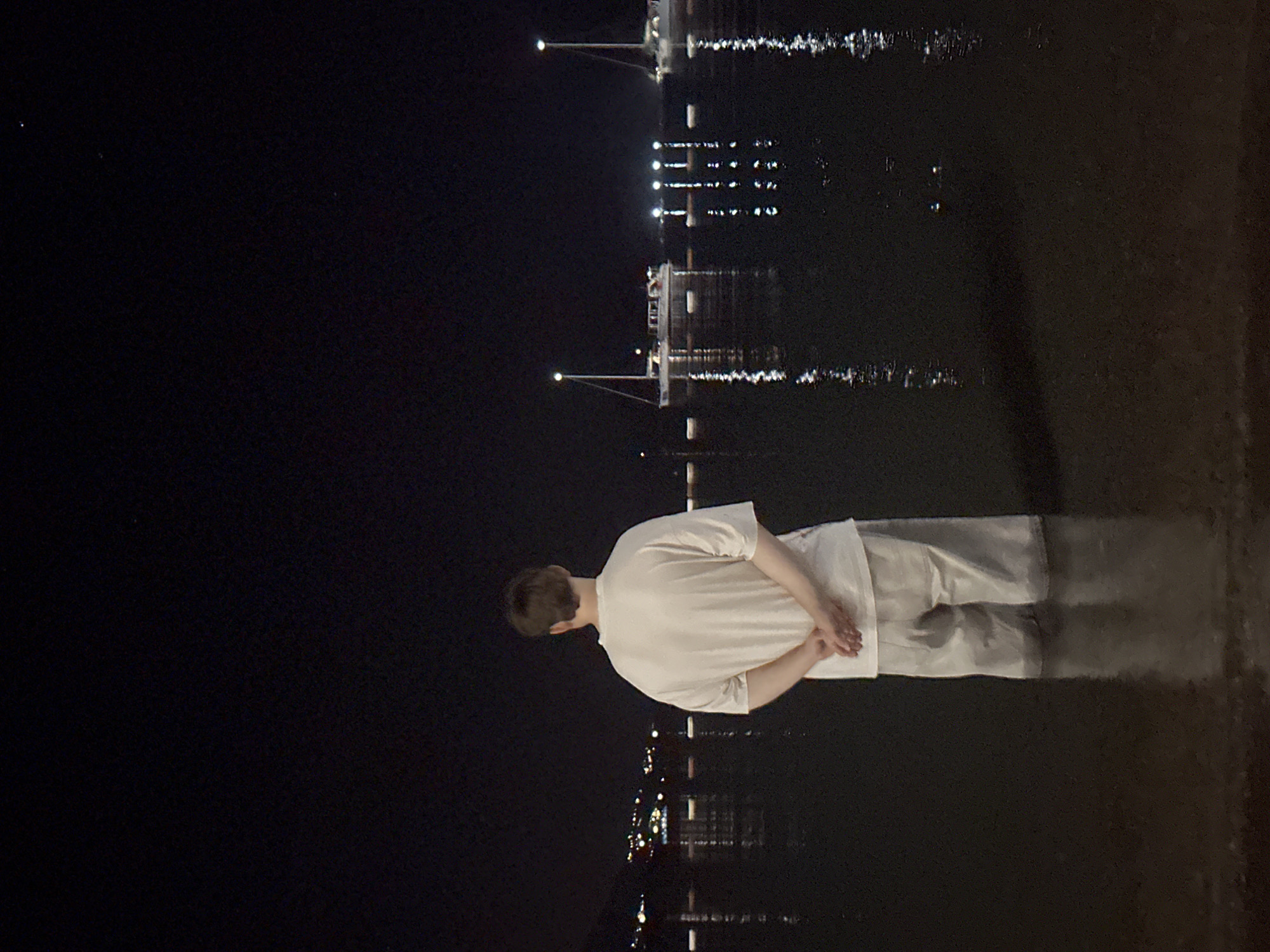 A lone individual stands in shallow water at night, facing away from the camera. The person is wearing a light-colored t-shirt and shorts, with their hands clasped behind their back. The water reflects the lights of a distant city skyline, which appears to be Budva, Montenegro. Several boats with illuminated masts are anchored in the bay, their lights also reflecting on the water's surface. The sky is dark, with a few faint stars visible.