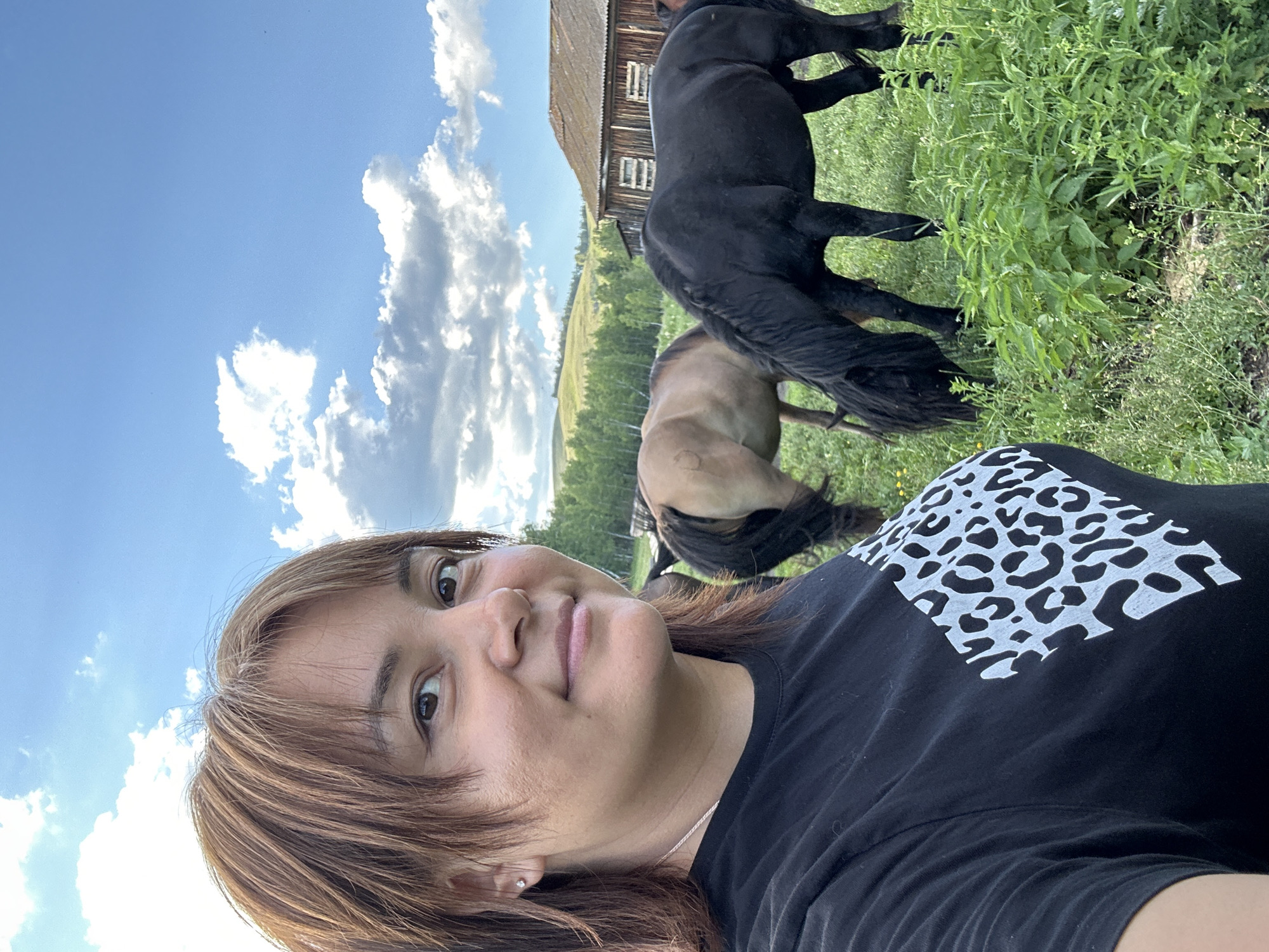 A woman is taking a selfie in a grassy field with two horses and a wooden building in the background. The woman has brown hair and is wearing a black t-shirt with a white leopard print design on the back. One horse is black and appears to be a Friesian, and the other is a lighter brown color. They are both grazing in the field. The wooden building looks like a barn or stable, and it is situated on a hill with trees behind it. The sky is blue with fluffy white clouds, suggesting it is a sunny day. The overall impression is a peaceful, rural setting. The image appears to have been taken in the late afternoon, as indicated by the angle of the sun. No visible text is present.