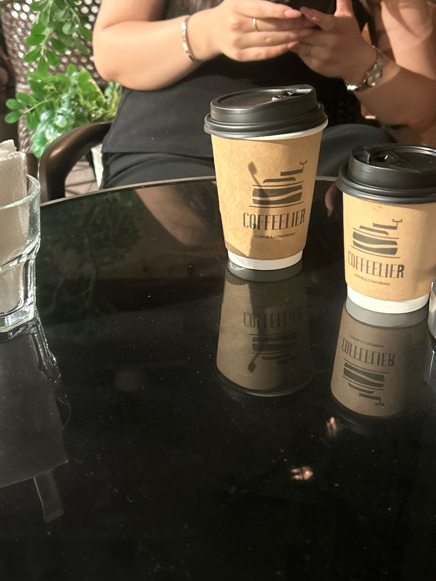 The image depicts a close-up view of two disposable coffee cups with black lids and brown sleeves, each featuring the logo "COFFEELIER" and smaller text "COFFEE & COWORKING". The cups are placed on a highly reflective black surface, likely a table, which shows a mirrored reflection of the cups. To the left of the cups, a portion of a clear glass container with a white object inside is visible. In the background, out of focus, a person's arm and hand can be seen holding a smartphone. A lush green plant is also partially visible in the upper left corner. The scene suggests a casual setting, possibly a cafe or coworking space, where someone is engaged with their phone while having coffee. The lighting is dim, creating a mood that is both intimate and slightly mysterious. There is no specific indication of the location being in Sumgayit, Azerbaijan.