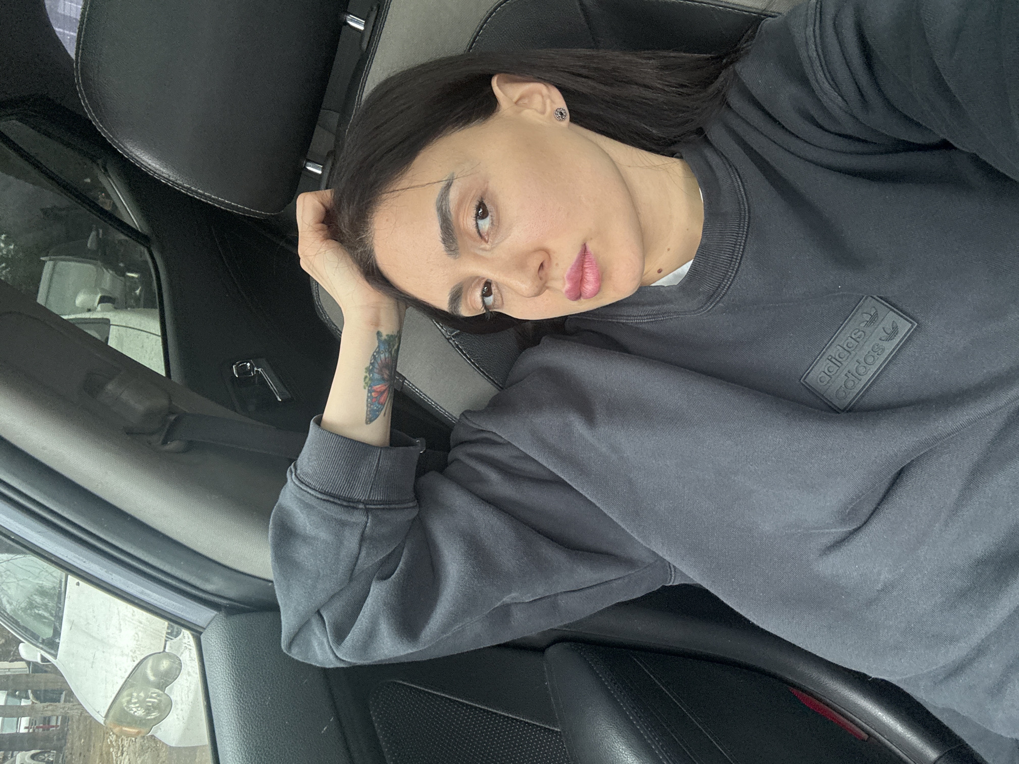 A female individual is seated in the passenger seat of a vehicle in what appears to be Tbilisi, Georgia. She has long, dark hair and is wearing a dark grey crew-neck sweatshirt with a raised "adidas" logo on the left chest, over a white undershirt. A colorful butterfly tattoo is visible on her right forearm. Her right hand rests on the crown of her head. She looks directly towards the camera, her lips adorned with light pink lipstick, and an earring is present in her left ear. The vehicle's interior features black leather seats with grey fabric inserts. Through the front passenger window, a light-colored minivan or SUV, appearing dirty, is parked parallel to the vehicle. The exterior background is indistinct, suggesting an urban or suburban setting under overcast conditions.