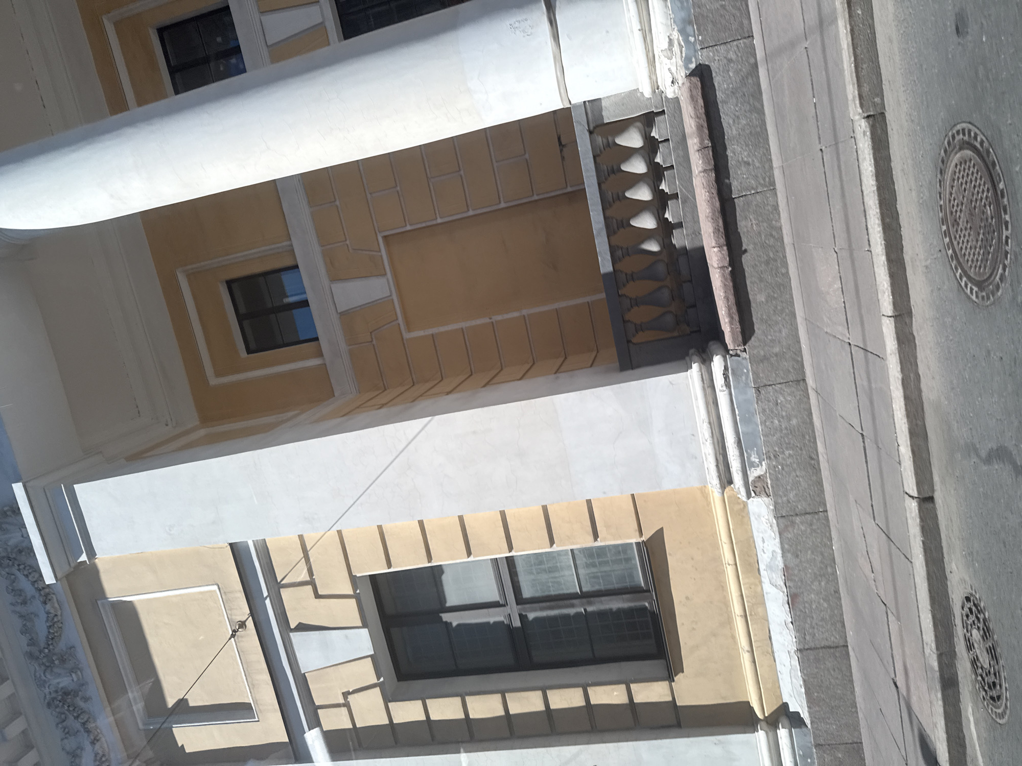 The image shows a view of a neoclassical building facade with large white columns and yellow walls. There are several windows with dark frames and grids. A decorative balustrade runs along a raised platform in front of some of the windows. The building is adjacent to a sidewalk made of stone slabs, which then meets a street with a manhole cover. The overall architectural style suggests it is an institutional or historical building, likely in Saint Petersburg, Russia, known for its abundance of neoclassical architecture. The lighting indicates it is daytime with bright sunlight casting distinct shadows. There are no people visible in the image.