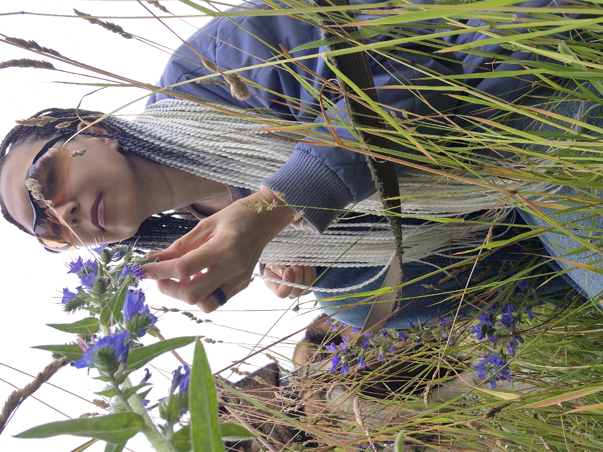 A medium shot captures a woman with long, braided hair, some of which is white, in an outdoor setting. She is wearing orange-tinted sunglasses, a blue jacket, and blue jeans. The woman is kneeling in tall grass and appears to be smelling a cluster of small purple flowers. Her right hand is near the flowers, and she is wearing a dark ring on her index finger. In the background, a dog, possibly a German Shepherd mix, is partially visible amongst the grass and flowers. The environment appears to be a meadow or field with a neutral sky. There is no discernible information within the image to place it in Chaikovsky, Russia.
