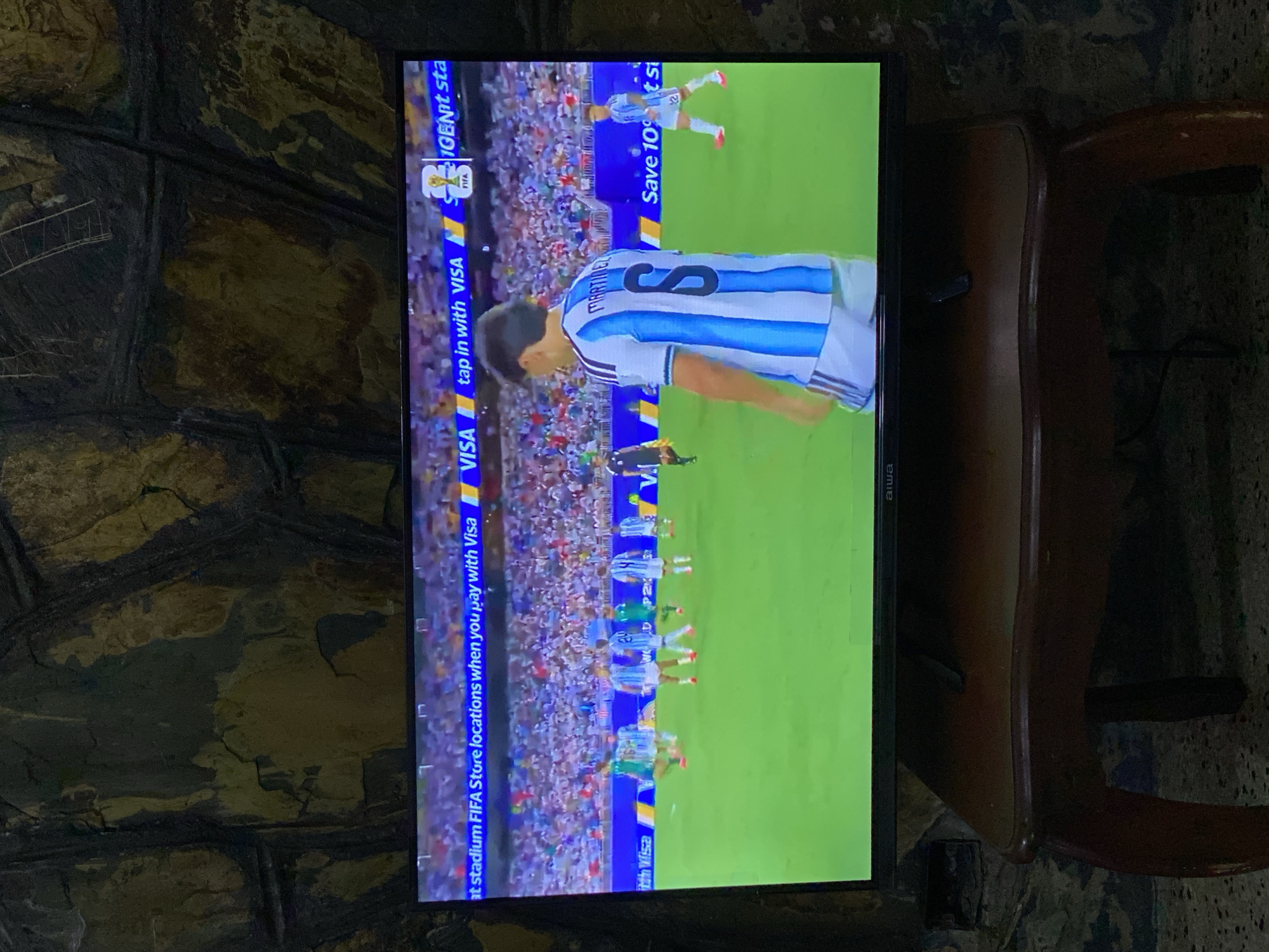 An indoor scene features a large television screen displaying a soccer match. The screen is positioned on a dark wooden table with curved legs, against a textured, stone-like wall. The soccer game shows players in blue and white striped uniforms on a green field, with spectators visible in the background. A banner along the bottom of the screen displays text related to VISA and FIFA. The lighting suggests the room is dimly lit, with the television screen being the primary light source. There are no visible indications placing this scene specifically in Tinaquillo, Venezuela; it appears to be a generic indoor setting.