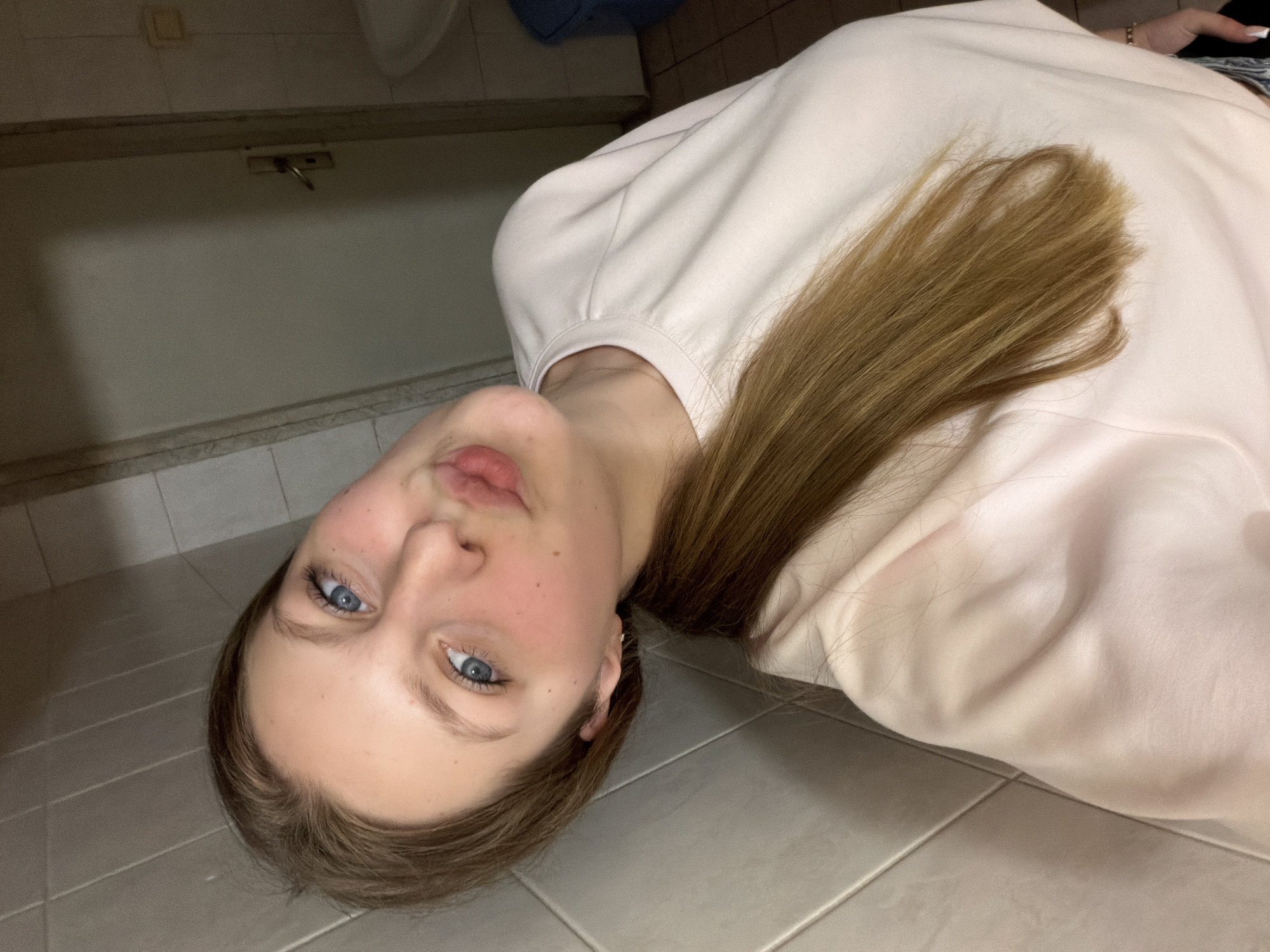 A young woman with blue eyes, light skin, and brown hair styled in a ponytail is taking a selfie in a tiled bathroom. She is wearing a light pink long-sleeved shirt. Her lips are pursed. The background shows a tiled wall, a door with a handle, and part of a sink and a blue trash bin. This scene is located in Burgas, Bulgaria.
