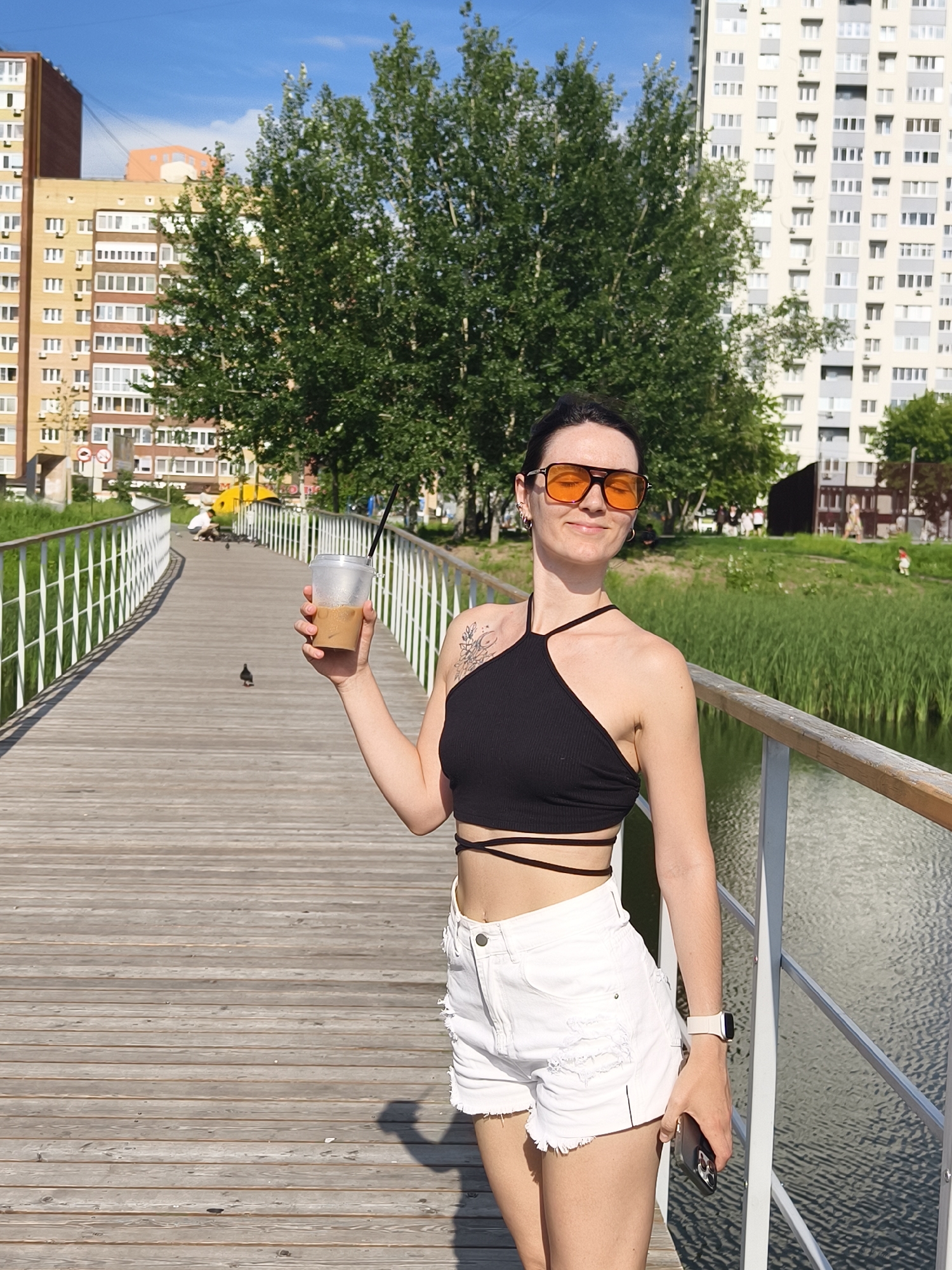 A medium shot captures a woman standing on a wooden bridge over a body of water, holding a plastic cup with a beverage. She is wearing a black halter top, white distressed denim shorts, and large orange-tinted sunglasses. Her right arm is extended, holding the cup, while her left hand rests near her shorts, holding a smartphone. A smartwatch is visible on her left wrist. The background features tall apartment buildings, trees, and green reeds along the water's edge, under a clear blue sky. The scene appears to be in Tyumen, Russia, possibly in a park or recreational area. There are pigeons on the bridge, and other people are visible in the distance.