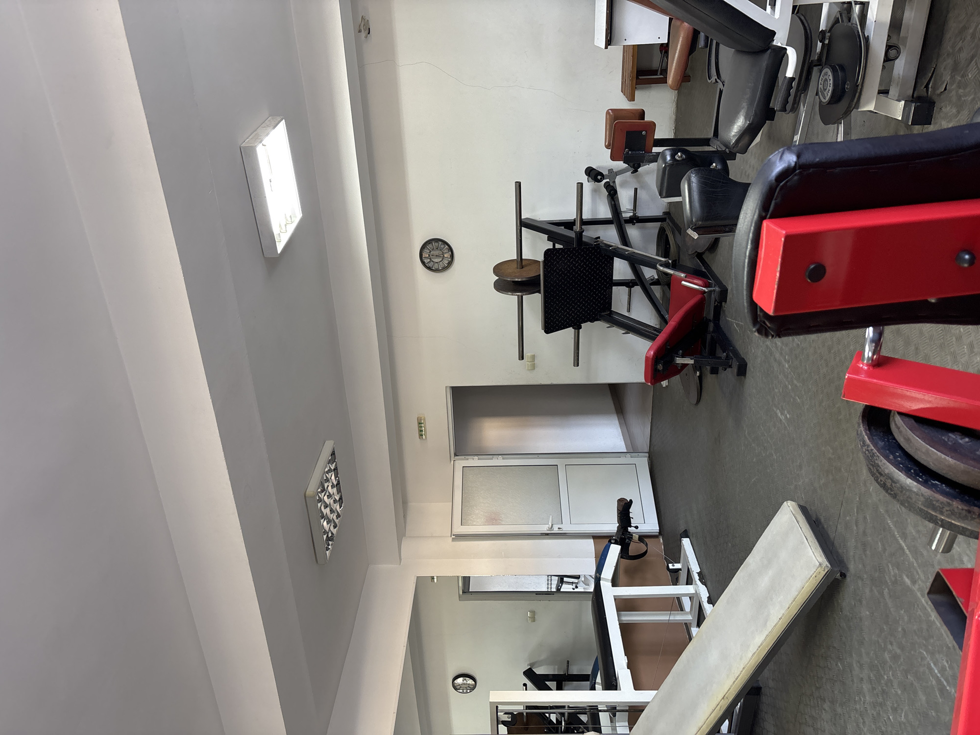 A photograph depicts the interior of what appears to be a gym. Weightlifting machines and benches are arranged on a dark gray floor. The walls are white and adorned with a clock. Lighting fixtures are mounted on the ceiling. A door is visible in the background. The location is identified as the city of Zlatograd, Bulgaria. There are no people present in the image.