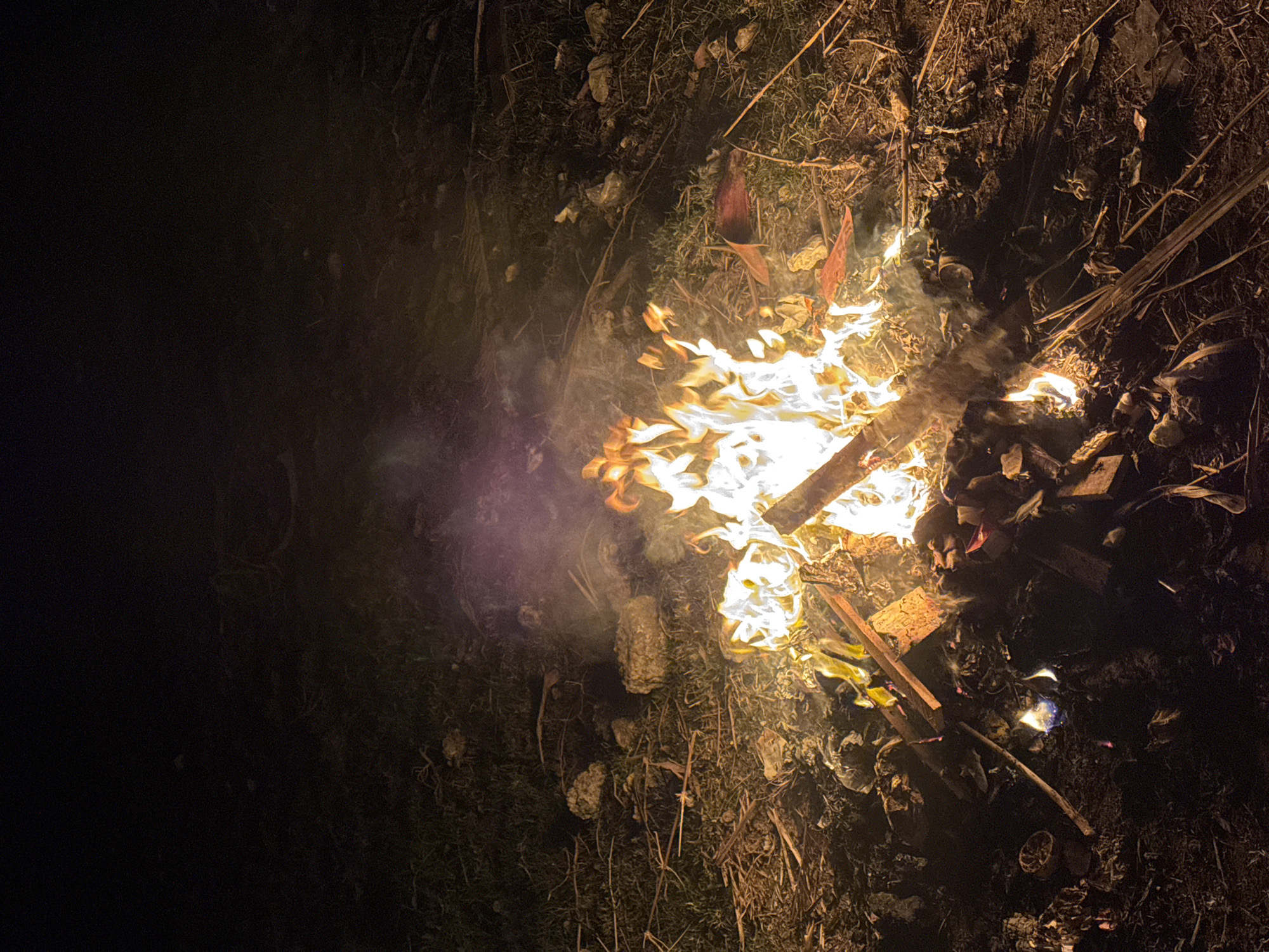 A nighttime scene depicts a bonfire in Kuta, Indonesia. The fire burns with bright orange and yellow flames, casting light on the surrounding dry grass and debris. Wisps of purple smoke rise from the flames, partially obscuring the dark background. Scattered twigs, leaves, and a few small stones are visible around the bonfire. The ground appears to be uneven and covered with organic matter. There are no people visible in the image. The lighting is dim, with the bonfire being the primary source of illumination.