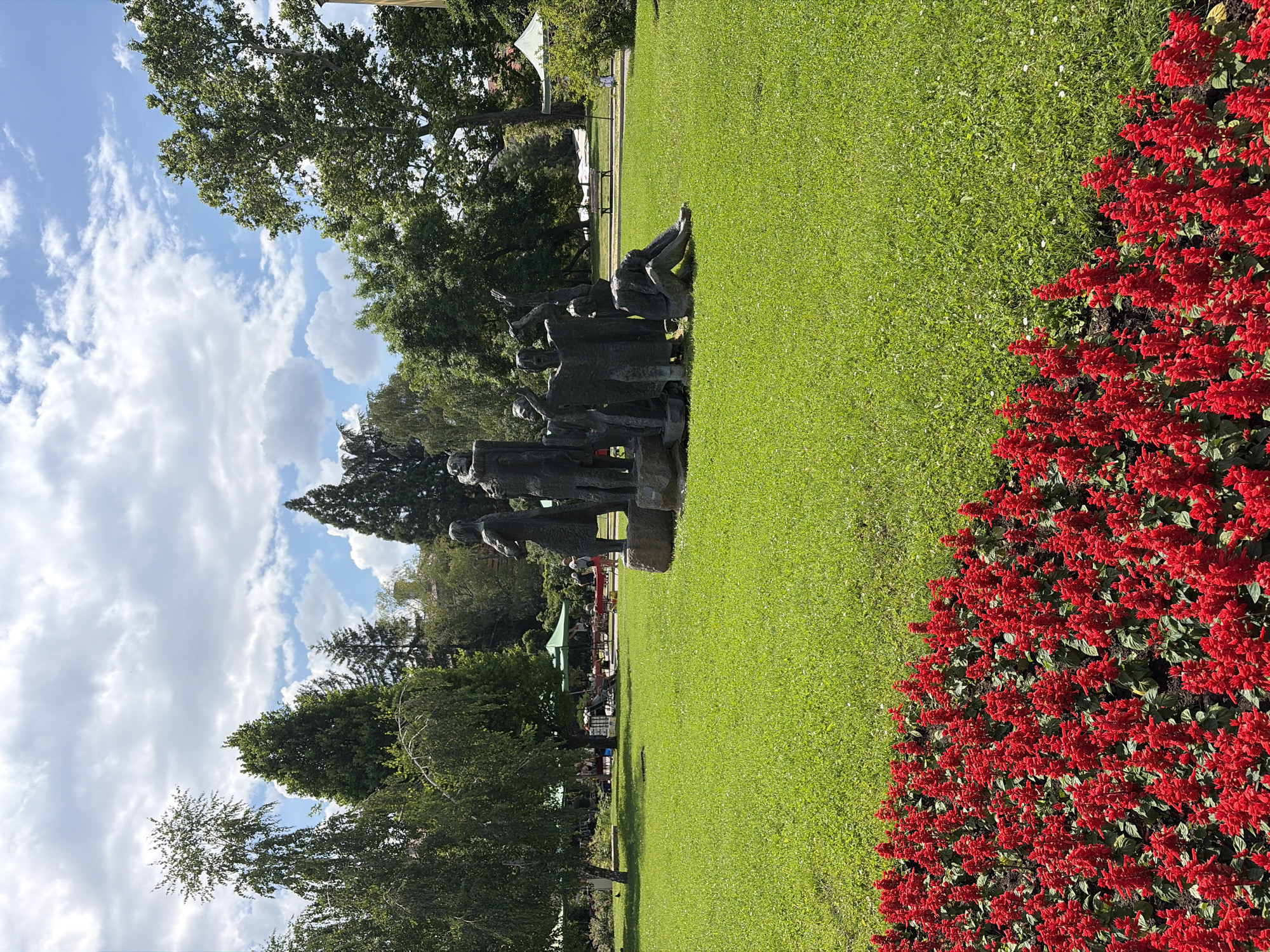 The image depicts a park scene on a bright, partly cloudy day. In the foreground, a bed of vibrant red flowers lines the bottom of the frame. Behind the flowers, a large expanse of lush green grass stretches across the middle ground. A bronze sculpture of several figures, some standing and one seated in distress, is centrally located on the lawn. Beyond the sculpture, more trees and greenery form the background, with a few tables and umbrellas suggesting a cafe or market area. In the distance, a white bench and path are visible. The overall impression is that of a well-maintained public space. This scene is located in the City Garden (Gradska Gradina) in Sofia, Bulgaria.