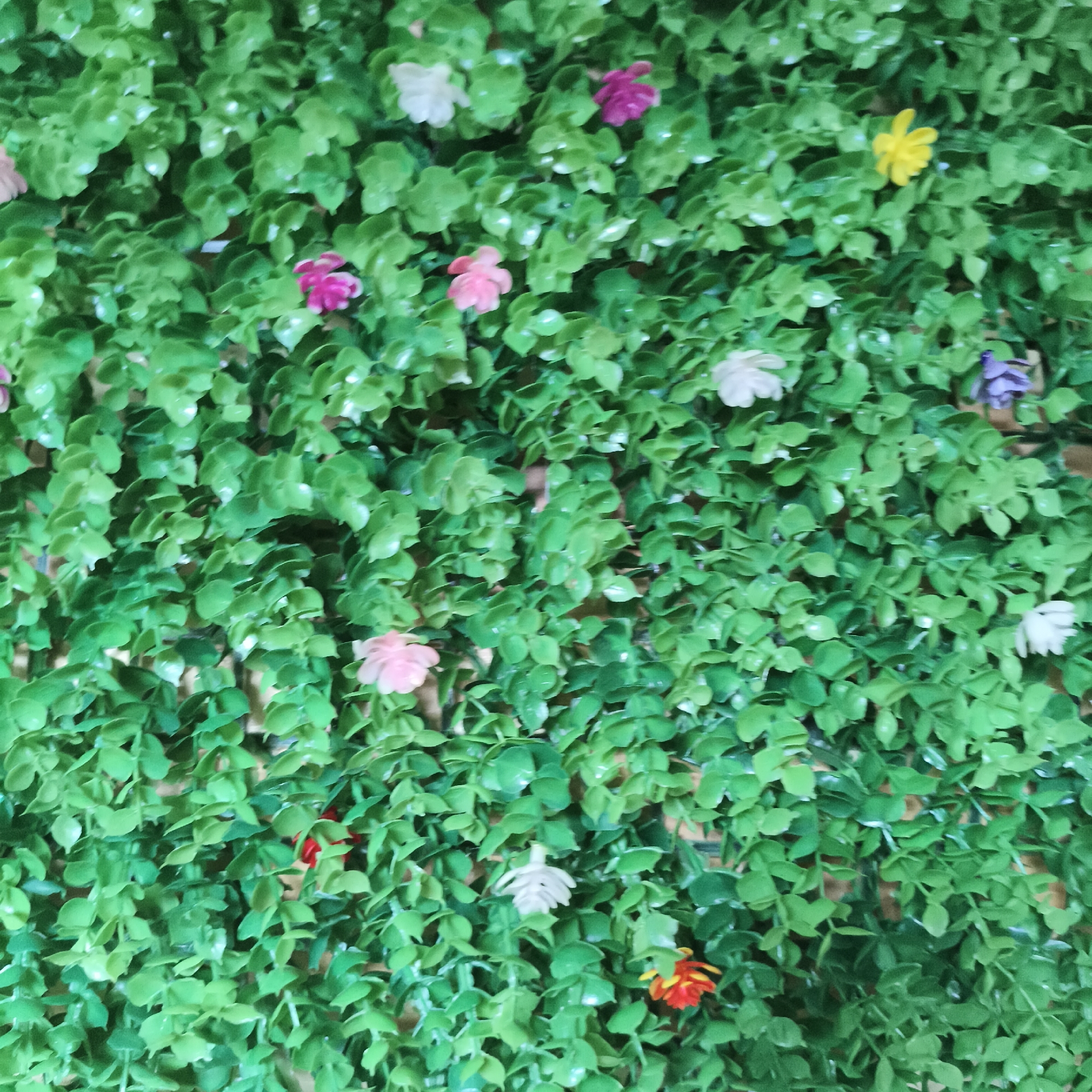 The image shows a wall covered in artificial green foliage with scattered small artificial flowers of various colors including pink, white, yellow, purple, and orange. The composition is dense, obscuring the wall behind the foliage. No people are visible. The location is not possible to determine from this image.