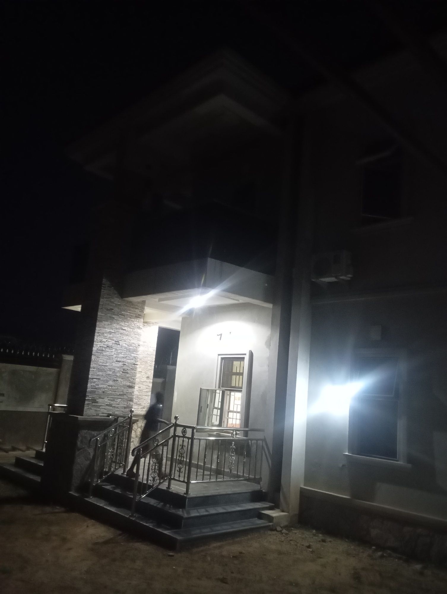 The image captures the exterior of a multi-story building at night, illuminated by artificial light sources. The structure features a light-colored, possibly stuccoed or painted, facade. A prominent entrance area includes a set of dark-colored steps with ornate metal railings, displaying both dark and reflective metallic elements, leading to a raised platform. To the left of the entrance, a section of the building's wall is faced with textured stone cladding in horizontal layers of varying grey and beige tones. A heavily blurred, indistinct human figure is partially visible on the steps or platform to the left of the primary entrance light, appearing in dark clothing. An exterior air conditioning unit is mounted on the upper right section of the building. Windows are visible, with one on the right side exhibiting a strong light flare. The ground in the immediate foreground is unpaved soil or dirt. The overall scene is dark, indicative of nighttime conditions, in the city of Unknown, Unknown.