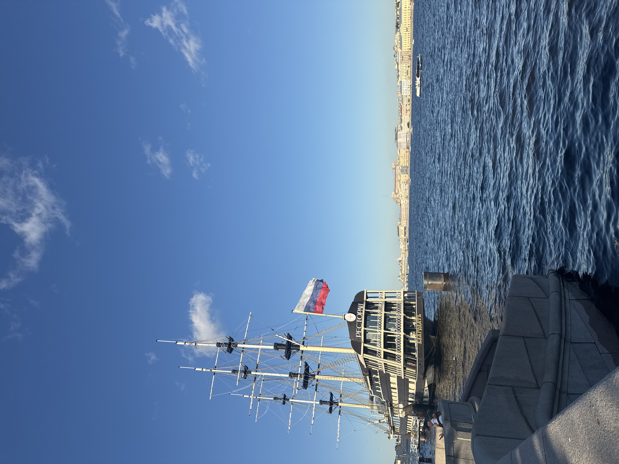 The image depicts a traditional sailing ship docked on the Neva River in Saint Petersburg, Russia. The ship, which appears to be converted into a restaurant as indicated by the Cyrillic text "РЕСТОРАН" on its stern, is moored alongside a stone embankment. A Russian flag flies from the stern. In the background, the cityscape of Saint Petersburg unfolds with a line of historic buildings under a clear blue sky with sparse white clouds. The water of the Neva is calm with gentle ripples. There are a few people visible sitting on the embankment in the foreground, and a small boat is seen in the distance on the river. The lighting suggests it is late afternoon or early evening.