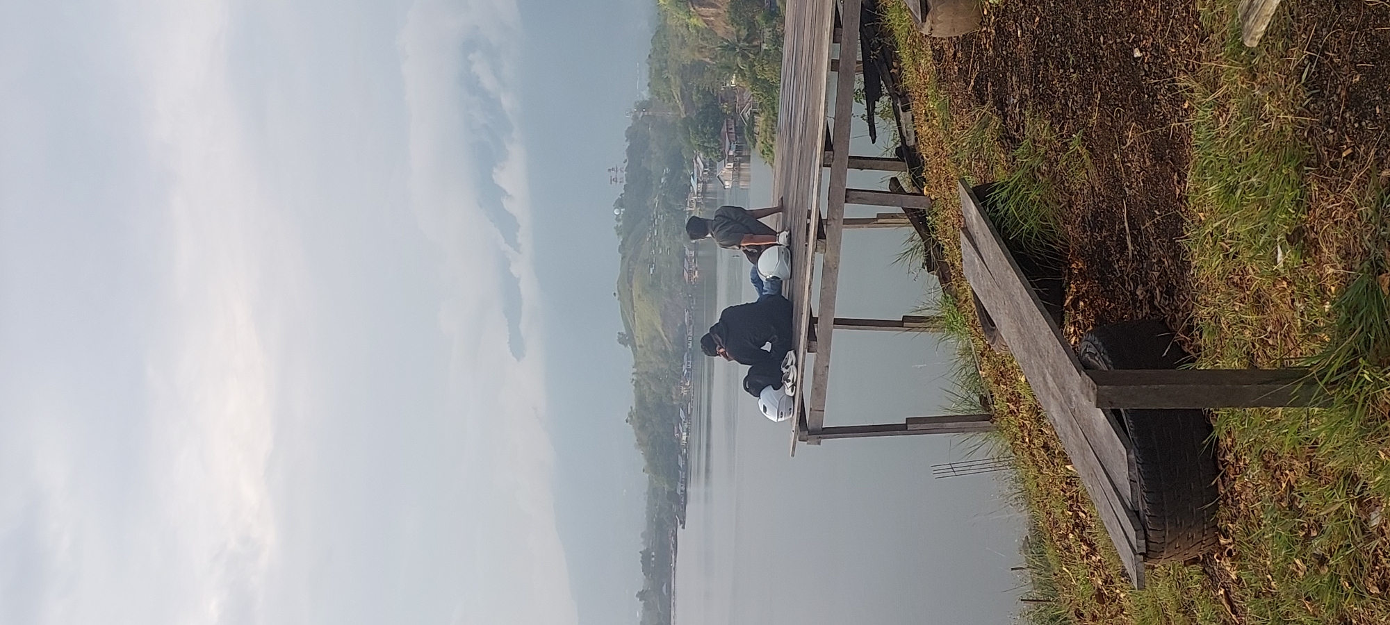 The image captures a serene landscape in Hobong, Indonesia, featuring two individuals seated on a wooden platform overlooking a vast body of water. The platform, constructed with weathered planks, extends over the water's edge, suggesting a vantage point for enjoying the scenery. On the platform, two helmets, a pair of shoes, and a bag are placed, indicating the presence of the two individuals who appear to be engaged in quiet contemplation or conversation.
The backdrop consists of rolling hills dotted with houses, some of which are built on stilts along the water's edge, characteristic of coastal or lakeside communities. The sky is overcast, with soft, diffused light casting a gentle glow over the scene. The foreground is characterized by dry foliage and grass, with a rough, wooden bench made from a tire and planks adding to the rustic charm of the location. The overall atmosphere is peaceful and tranquil, inviting a sense of calm.