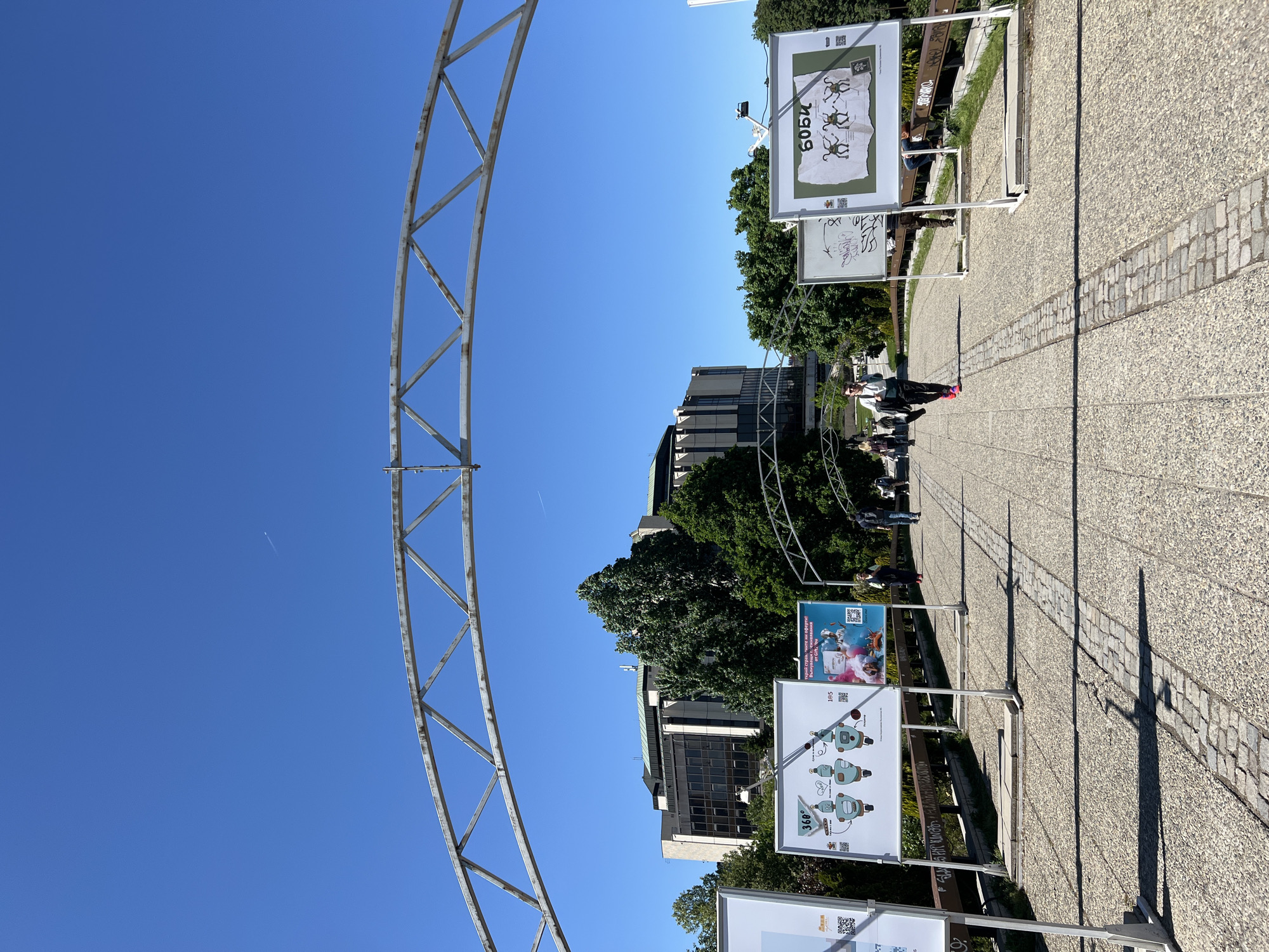 The image shows a pedestrian walkway in Sofia, Bulgaria, lined with exhibition panels. A metal archway frames the view. People are strolling along the path. Buildings and trees are visible in the background under a clear blue sky. The walkway is paved with light-colored aggregate and features a brick-lined section. Shadows of the panels and archway are cast on the walkway. The exhibition panels contain informational posters.
