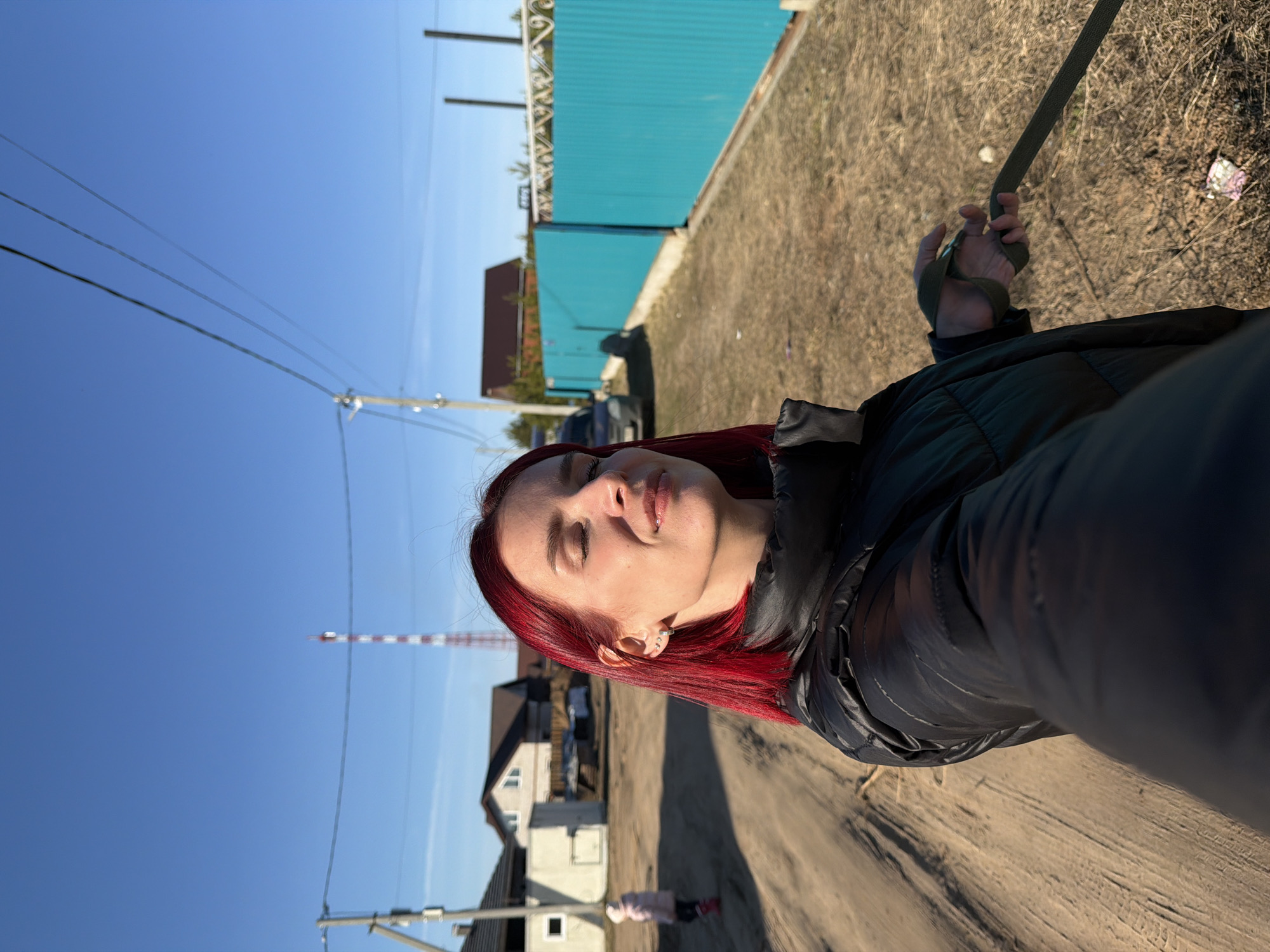 The image, when viewed correctly oriented, features a young woman with vibrant red hair, facing slightly left with her eyes closed or squinting, bathed in bright sunlight. She wears a dark, possibly black, puffer jacket. Her right hand is visible in the foreground, holding a dark green strap or leash, suggesting she might be walking an animal or carrying an item.
The setting appears to be a rural or suburban area in Chaikovsky, Russia, under a clear, bright blue sky with no clouds. The ground is a dirt or gravel path with sparse dry grass and weeds. To the left, utility poles with power lines line the path, and a house with a white base and a dark roof is visible. Further in the distance on the path, a small, indistinct figure (possibly a child) can be seen. On the right side, a prominent bright turquoise fence or wall runs along the path, behind which a car and another house with a brown roof are partially visible. In the background, a red and white striped tower stands against the horizon, likely a communication or water tower.
The strong, direct lighting indicates it is midday or early afternoon. The woman's relaxed expression suggests she is enjoying the sunny weather. There is no visible text in the image.
