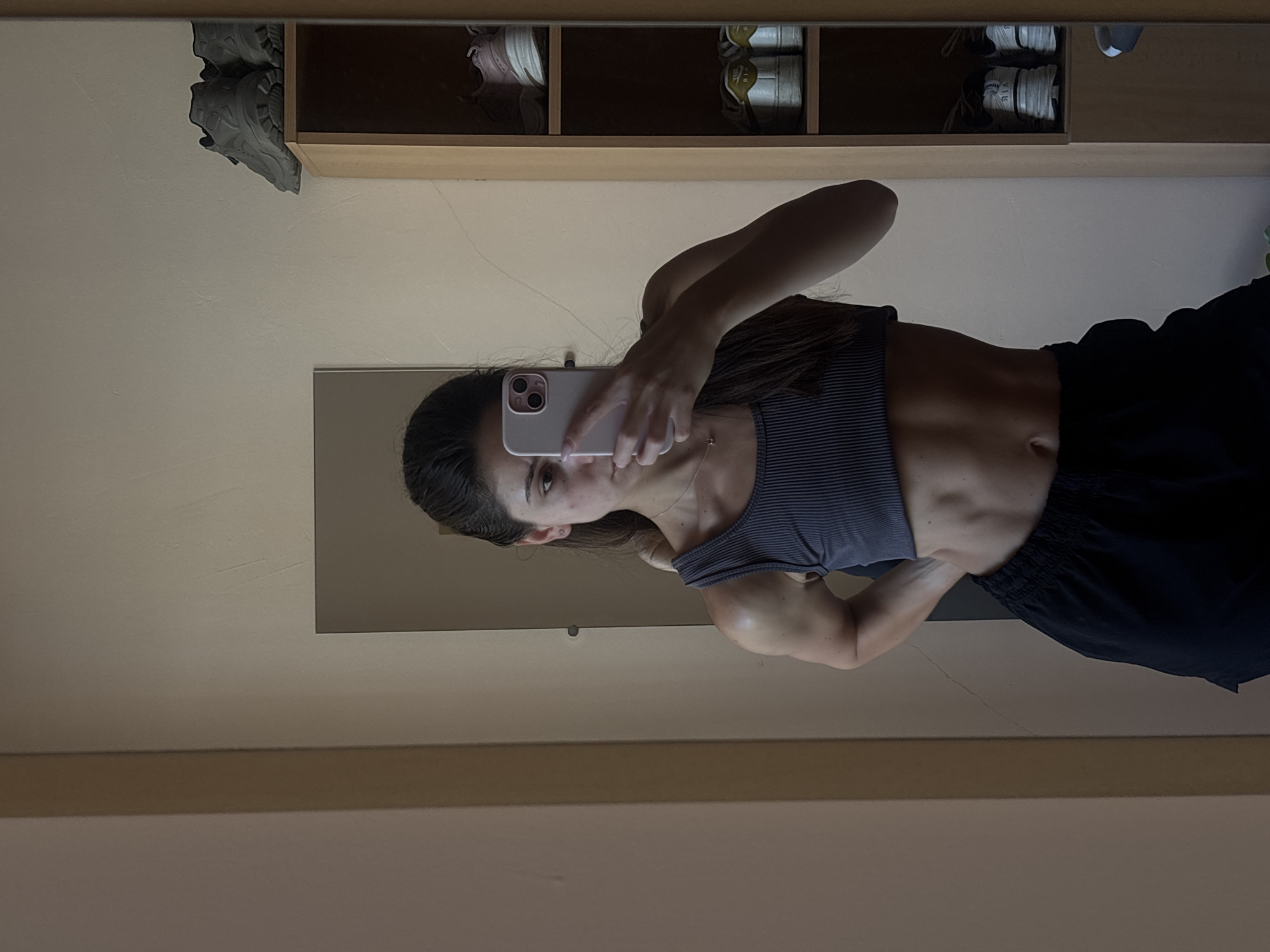 In Varna, Bulgaria, a young woman with dark hair, pulled back, is taking a mirror selfie. She is wearing a dark grey ribbed tank top and black shorts. Her toned physique is visible, with defined abdominal muscles and arms. She is holding a smartphone in her right hand, angled to capture her reflection. To the right of the mirror, a tall wooden shelf unit displays several pairs of athletic shoes. The background appears to be an indoor space with neutral-colored walls.