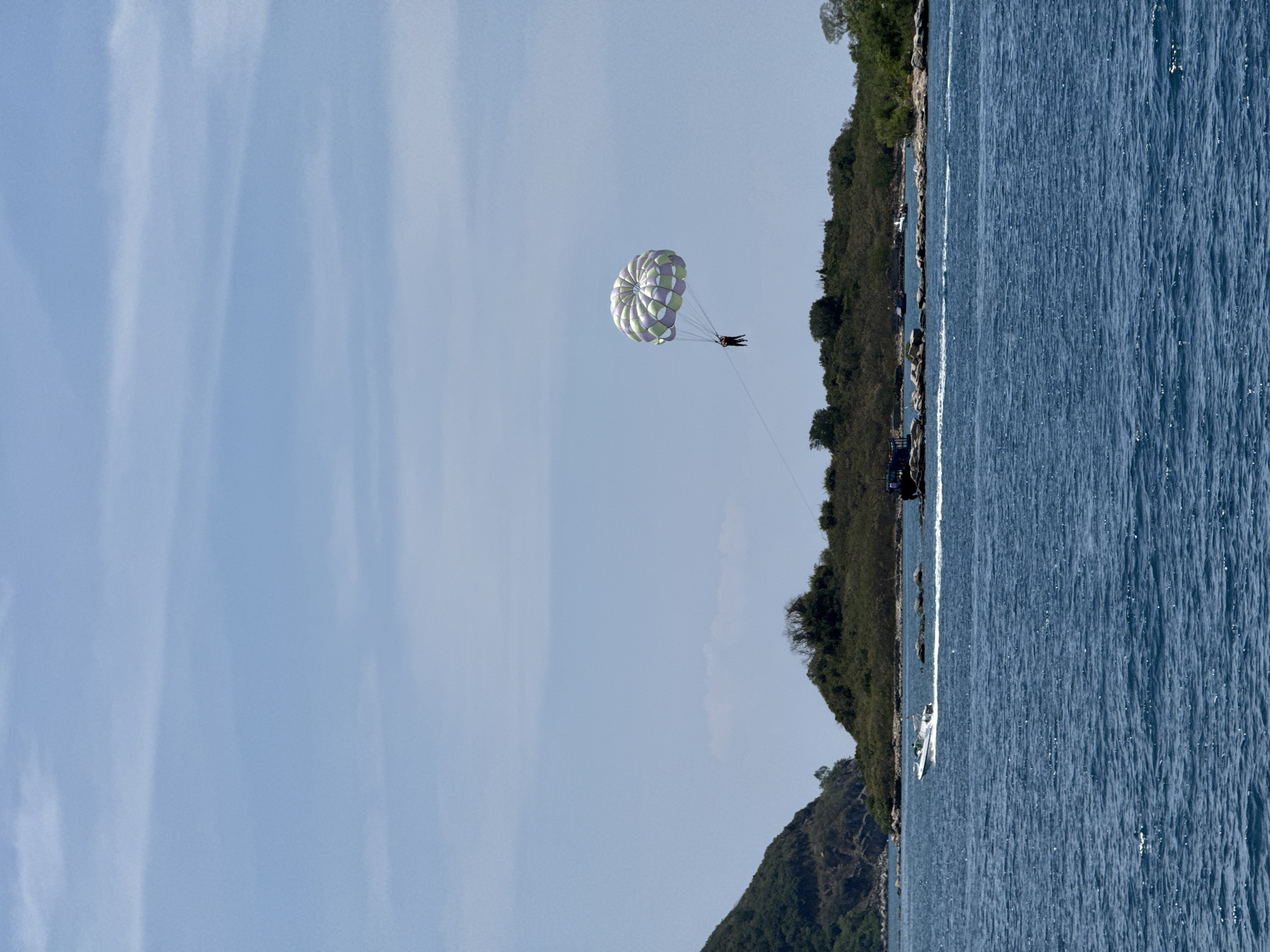 A parasail is suspended mid-air above the ocean near the coast of Nha Trang Ward, Vietnam. The parachute is white with lavender and green stripes. Two people are being towed behind a boat, which is visible in the ocean. The landscape includes blue ocean and sky. The coastline is lush with green vegetation.