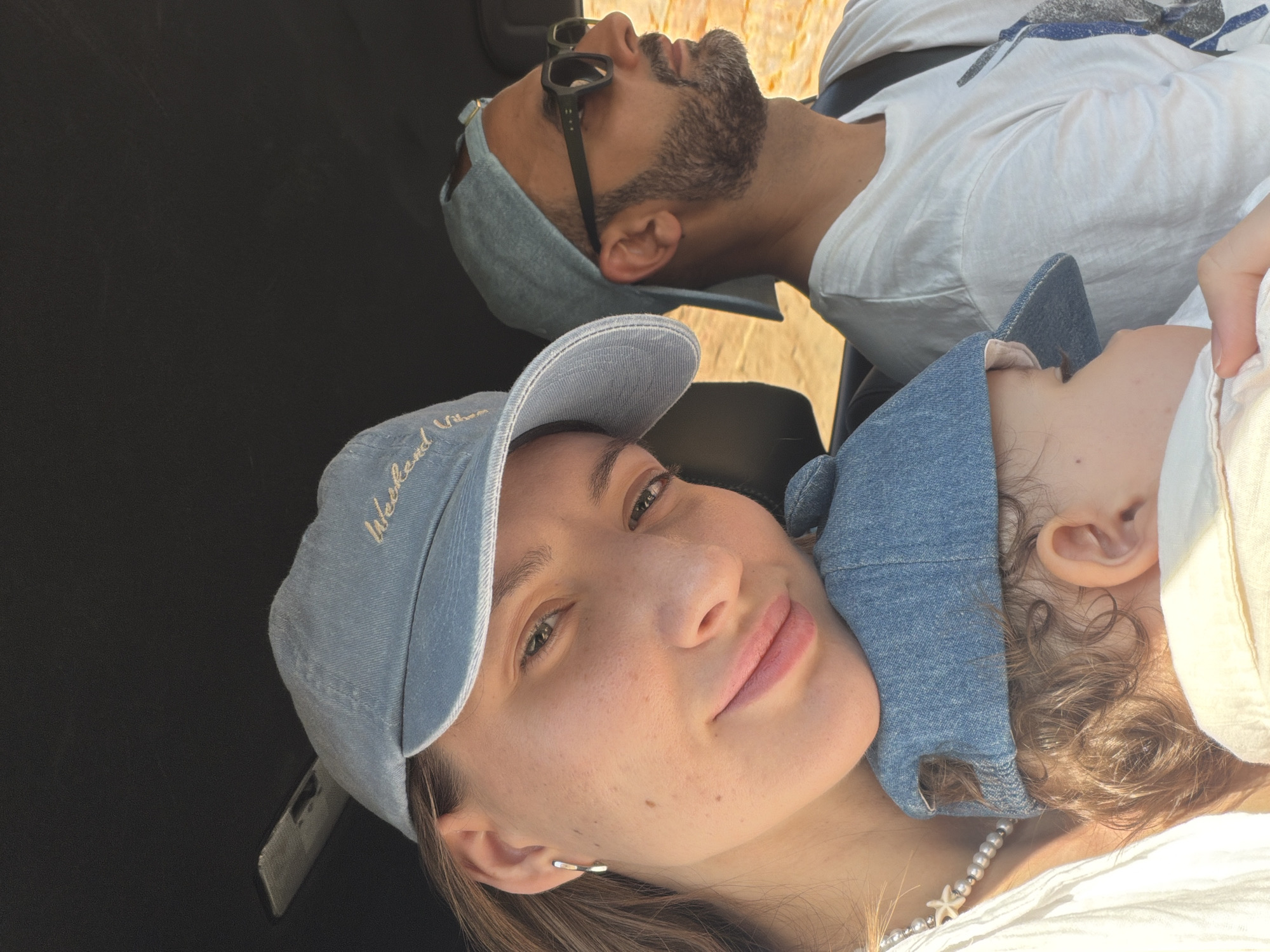 This is a close-up, selfie-style photograph taken inside a vehicle in Sharm El Sheikh, Egypt. The primary subjects are a woman in the foreground, a baby held by the woman, and a man in the background driving the vehicle. The woman, wearing a light blue denim baseball cap and a white shirt with a pearl necklace, is looking at the camera with a slight smile. Her hair is brown and visible on her left side. The baby, also wearing a light blue denim cap, is nestled against the woman and appears to be asleep, with its head and part of its face visible. The man, wearing a white t-shirt, a light blue denim baseball cap, and dark sunglasses, is looking forward, presumably at the road. He has a beard. The interior of the vehicle, including a dark headliner and a seatbelt, is visible. The background outside the vehicle shows a blurred desert landscape, consistent with the environment of Sharm El Sheikh. The lighting suggests it is daytime, with sunlight illuminating the scene.