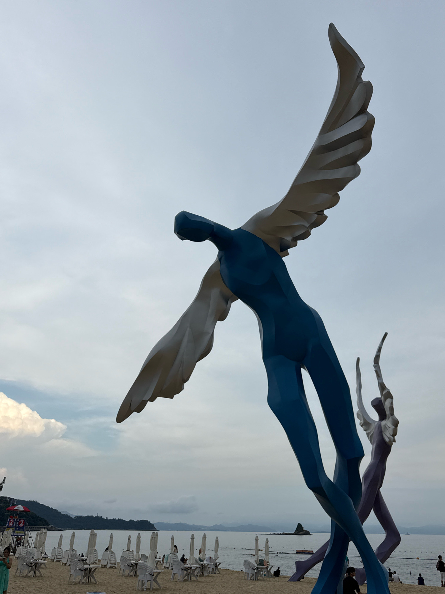 This is a low-angle, full shot of a large, abstract sculpture of figures with wings on a beach. The main sculpture is of a stylized, blue human figure with large, white wings. To the right, a smaller, purple figure with white wings is also visible. The sculptures are positioned on a sandy beach with rows of white chairs and umbrellas in the background. The ocean is visible beyond the beach, and a distant mountainous coastline is on the horizon. The sky is overcast. In the foreground, parts of the sculptures' bases are visible, along with some beachgoers. The sculpture is located in the Yantian District of China.