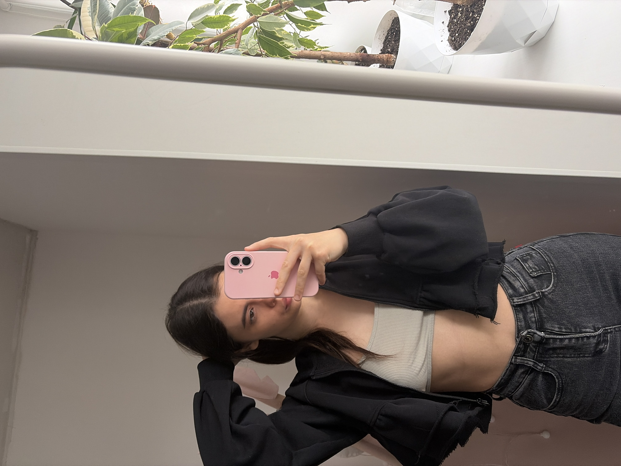 A young woman is captured in a mirror selfie, with the image rotated 90 degrees to the left. She holds a light pink smartphone, possibly an iPhone given the visible Apple logo and dual camera lenses, directly in front of her face, obscuring part of her features. Her dark hair is parted in the middle and falls around her shoulders. She wears a black zip-up jacket over a light cream or white ribbed crop top, exposing her midriff, and dark grey high-waisted jeans. Her left arm is bent, and her hand rests near her head.
The background, visible in the mirror's reflection, shows an indoor setting. Above the woman's head, a white shelf or railing spans horizontally. Behind this, several potted plants with variegated green and white leaves are visible, suggesting a window sill or a similar elevated space. The plants are in white, geometrically patterned pots, some filled with dark soil. The lighting is even, indicating an indoor environment, possibly during the daytime, but specific time or weather cannot be determined. The woman's expression is neutral as she looks towards the phone. The scene is casual and appears to be in a domestic setting, located in Chaikovsky, Russia.