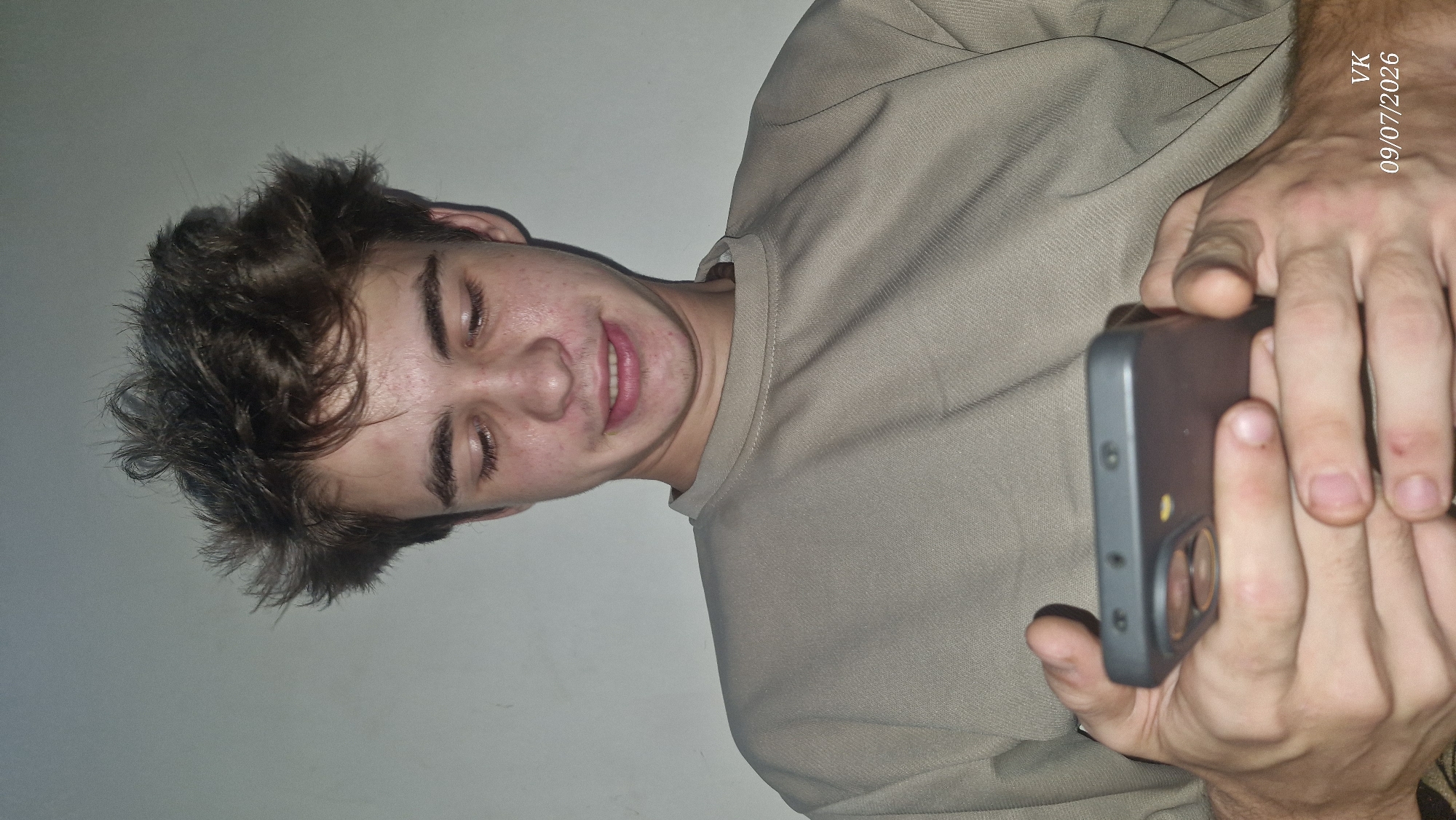 A young man with messy brown hair and fair skin is looking down at his smartphone. He is wearing a beige crewneck shirt. His eyes are closed, and he has a slight smile on his face. The background is a plain, light-colored wall. The image includes a watermark in the bottom right corner that reads "VK 09/07/2026". The image does not show any elements that would indicate its location within the city of Vrindavan, India.