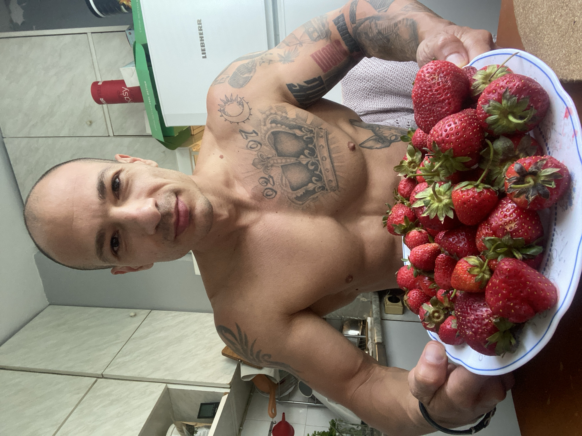 A shirtless man with visible tattoos on his chest and arms is holding a plate of strawberries in a kitchen. The man has a shaved head and is looking directly at the camera with a neutral expression. The kitchen appears to be somewhat cluttered, with cabinets and a refrigerator visible in the background. The strawberries are red and appear to be fresh. The image does not provide any information about the city of Pleven, Bulgaria.