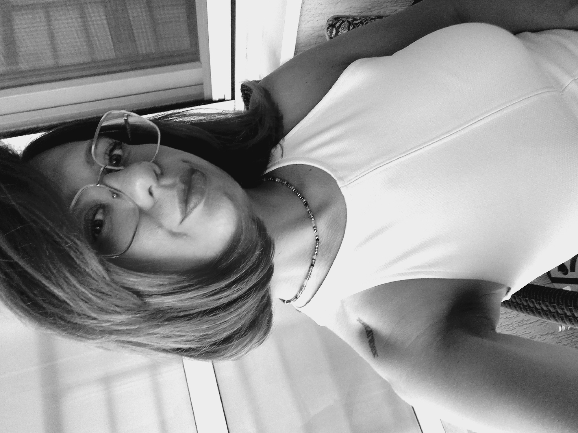 The black and white, high-angle, medium shot shows a woman from the chest up, facing the camera. She is indoors, by a window with vertical blinds. The woman has short, dark hair, and is wearing glasses with thin, wire frames. She has a beaded necklace on, and a white, sleeveless top with a seam running down the center. A small tattoo of Roman numerals is visible on her left shoulder. The background is blurred, showing a window on the right side and a wall on the left. The scene is a domestic setting, possibly a balcony or a room in an apartment. The lighting is soft, creating a gentle glow on the subject.