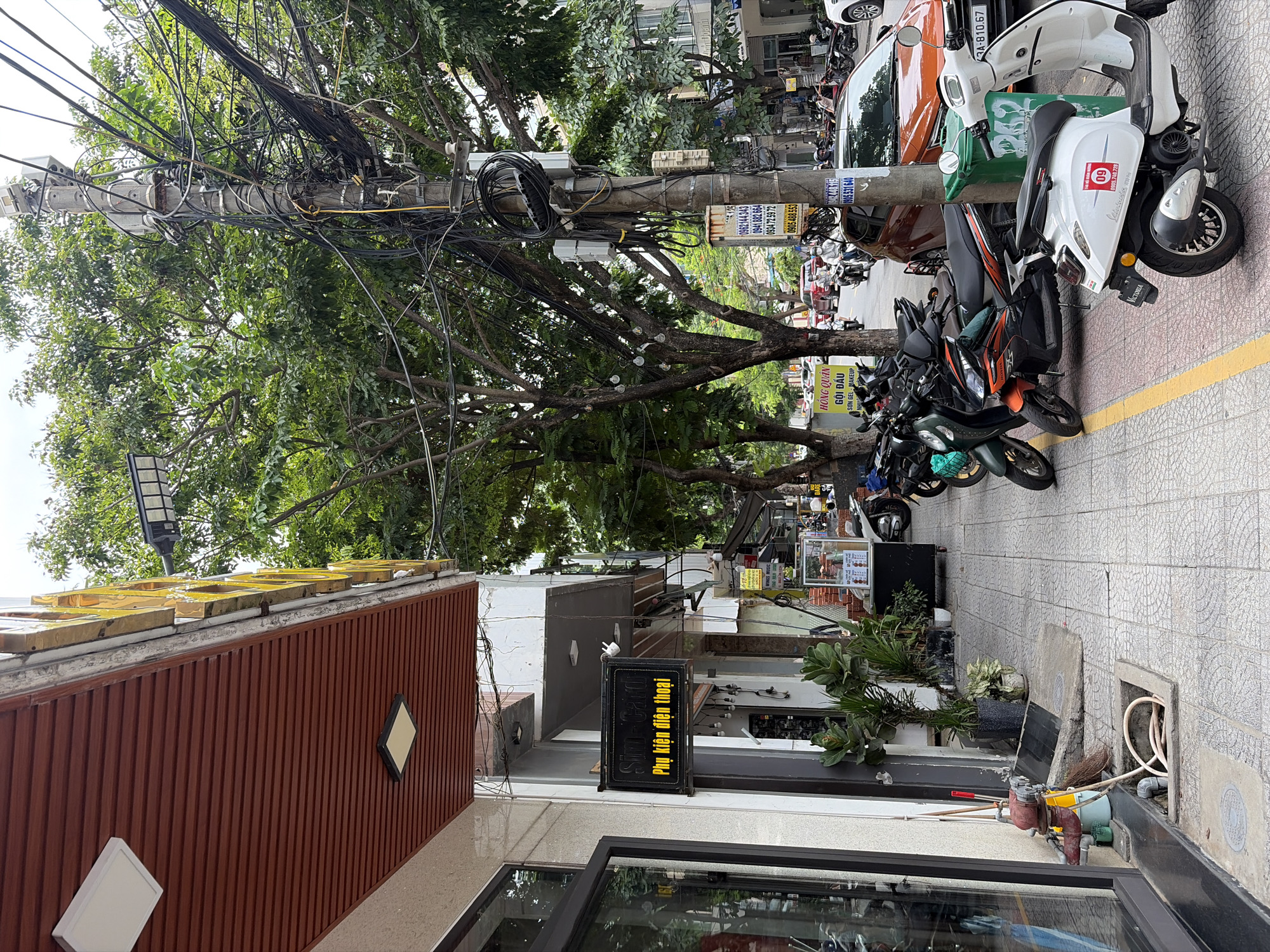 The image captures a street scene in Da Nang, Vietnam, focusing on the sidewalk and road in front of a row of shops. The architecture suggests a tropical climate with the presence of lush green trees lining the street and a visible overhead electrical grid. On the sidewalk, various items are present: a fire hydrant with a hose, planters with greenery, and several parked motorbikes, which are a common mode of transportation in Vietnam. One shop displays a sign that reads "Sim-Card" and "Phụ kiện điện thoại" (phone accessories) in yellow lettering against a black background. Further down the street, other shops and buildings are visible, along with more parked vehicles and pedestrians. The road appears to be a typical urban street with traffic, including cars and more motorbikes. The overall impression is one of a bustling commercial area on a cloudy day.
