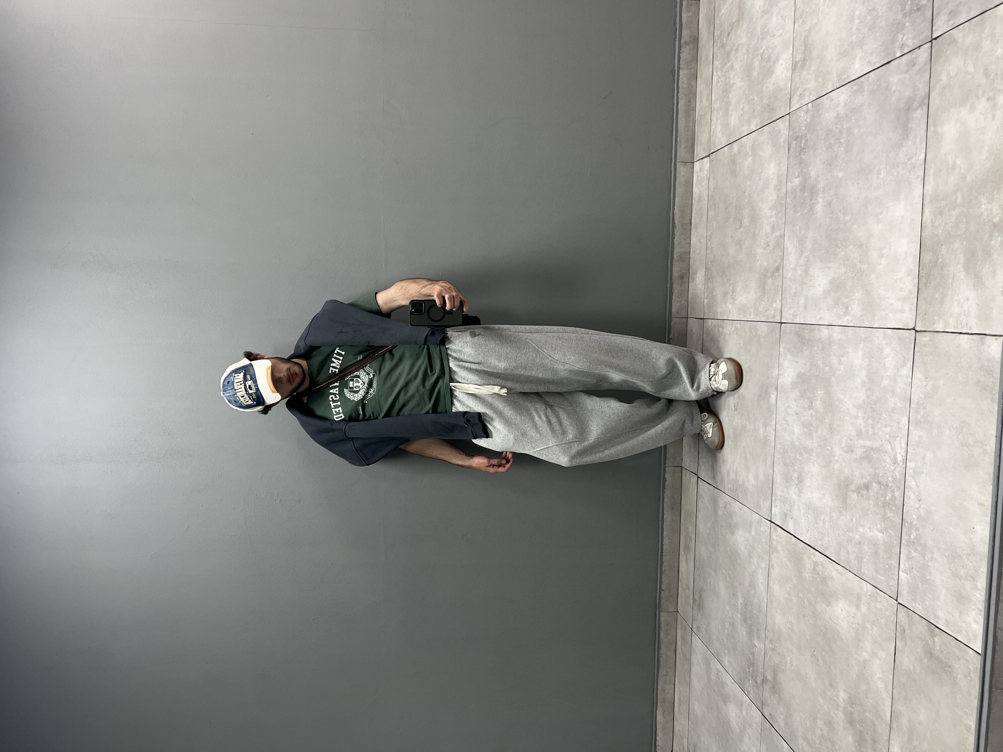 A person wearing a hat, t-shirt, hoodie, and sweatpants stands against a gray wall and gray tiled floor, holding a smartphone in their hand. The location is potentially in Yerevan, Armenia.