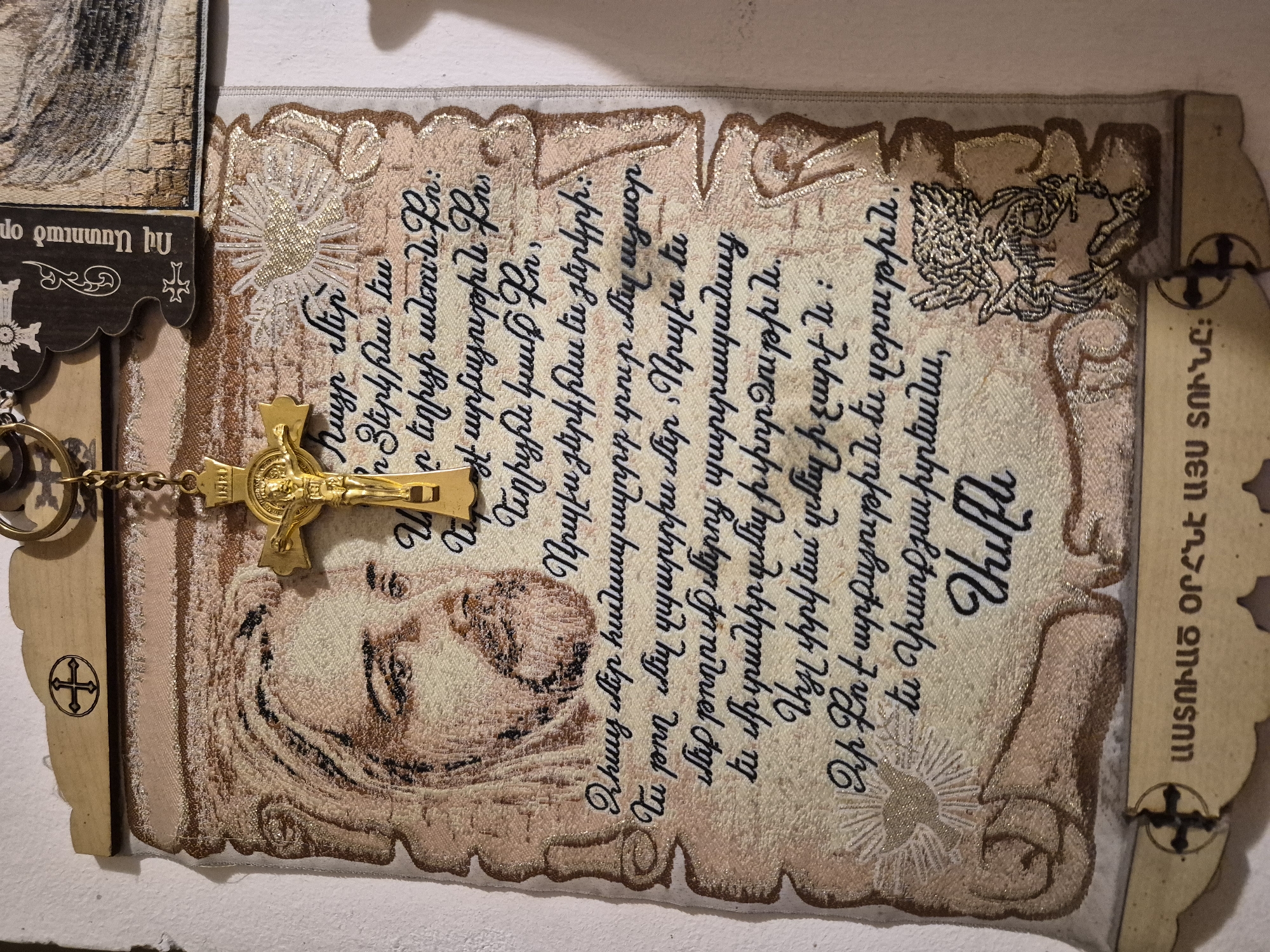 The image displays a vertically oriented embroidered religious textile, likely a wall hanging, adorned with a golden crucifix. The textile itself is primarily cream and beige, embellished with intricate gold and white embroidery, giving it a rich, textural appearance. A central embroidered portrait, possibly depicting a religious icon such as the Virgin Mary or a saint, with a solemn expression, occupies the lower portion. Above this figure, on the left, a white and gold embroidered dove, symbolizing the Holy Spirit, is prominently featured.
A golden crucifix, bearing the figure of Christ with "INRI" inscribed above, hangs from a delicate chain, overlapping the upper left section of the textile. The chain is attached to a small, dark wooden piece at the top left, which displays partially visible Armenian text and a cross symbol.
The main body of the textile is embroidered with legible Armenian script, identified as the Lord's Prayer ("Հայր մեր" - Our Father). The elegant cursive script is rendered in black thread, set against the pale background. The textile also features decorative gold-embroidered borders, designed to resemble an antique scroll or parchment.
On the right side of the image, a vertical wooden plaque is visible. It bears the Armenian inscription "ԱՍՏՈՒԱԾ ՕՐՀՆԷ ԱՅՍ ՏՈՒՆԸ։" (God Bless This Home), accompanied by a cross symbol. Another wooden piece, similar in style, is partially visible on the far left.
The scene conveys a sense of profound religious devotion, with the items appearing to be new or well-preserved, suggesting their purpose as devotional objects or gifts. The setting is indoors, characteristic of a home, church, or religious goods establishment in Yerevan, Armenia, as indicated by the Armenian language and religious motifs.