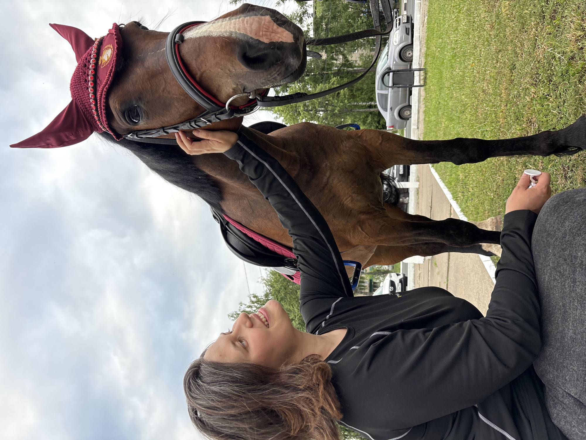 A woman with shoulder-length, wavy brown hair with streaks of grey smiles as she kneels next to a brown horse with a maroon fly bonnet adorned with red jewels. The horse wears a bridle with red trim. The woman is wearing a black long-sleeved shirt with white stitching and grey sweatpants. She is touching the horse's face with her right hand. In her left hand, she holds a small white object. The horse is standing on a grassy area next to a paved path. In the background, there are trees, cars, and buildings, indicating an urban setting. The sky is overcast.
