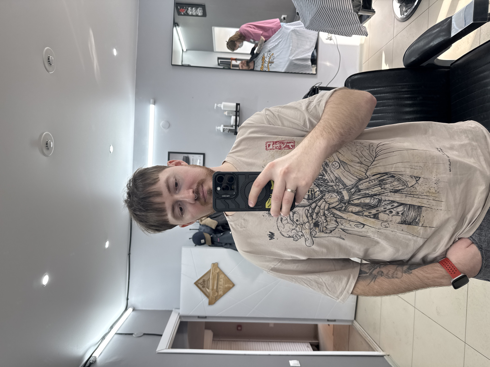 This image depicts the interior of a barbershop located in Votkinsk, Russia.
In the foreground, a male subject with light brown hair, a short fringe, a light beard, and a septum piercing is taking a mirror selfie with a black smartphone held in his right hand. He wears a beige t-shirt with a dark graphic design, a silver-colored ring on his left ring finger, a red smartwatch on his left wrist, and has a tattoo visible on his left forearm. His left hand is in his pants pocket.
The room features light grey walls and a white ceiling. Illumination is provided by multiple recessed spotlights and horizontal fluorescent tube lights mounted on the walls. Two white circular smoke detectors are affixed to the ceiling, and visible electrical conduits run along the walls and ceiling.
To the left of the primary subject, a white door with a geometric pattern is open, revealing a hallway with light-colored tiled flooring and a red fire alarm call point. A wooden, diamond-shaped sign featuring a car outline and the word "GARAGE" is mounted on this door. A coat rack with a dark jacket and a baseball cap is visible behind the subject.
To the right, a large mirror reflects a female barber, wearing a pink top, blue jeans, black gloves, and a face mask, attending to a male client seated in a black barber chair. The client is covered by a striped barber cape. Another black barber chair draped with a striped cape is partially visible in the right foreground. A small black wall shelf holds various bottles, and decorative elements are visible on the far wall within the mirror's reflection. The floor throughout the visible area is covered with light-colored tiles.