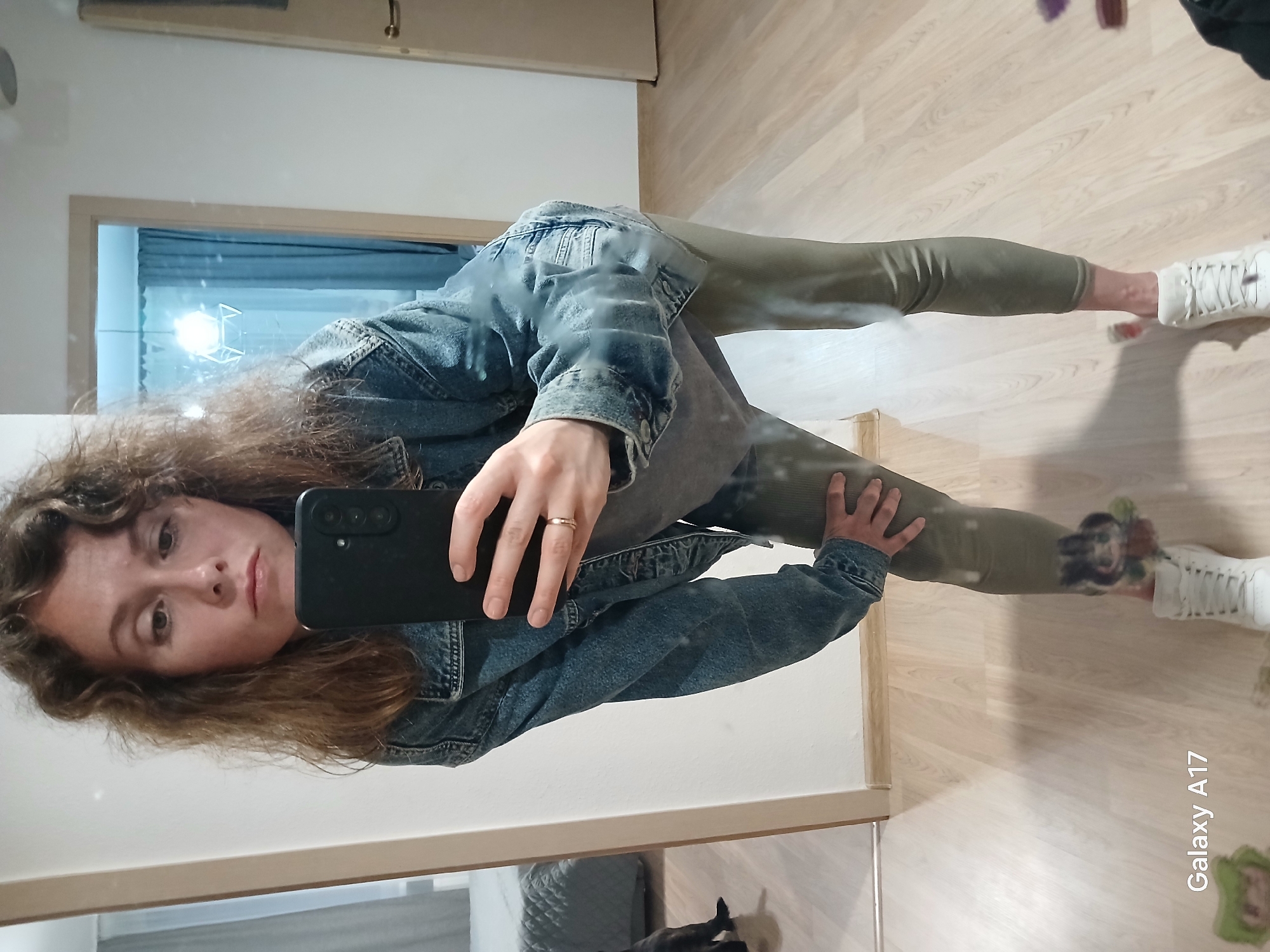 A woman is taking a selfie in a mirror. She is wearing a denim jacket and olive green leggings with white sneakers. The location appears to be indoors, likely a residence or apartment. The background is a wooden floor, white walls and a door. The mirror reflects a partially visible gray quilted bag on the floor. The writing "Galaxy A17" can be seen in the bottom right of the image. There is no evidence to indicate this image was taken in Yekaterinburg, Russia.