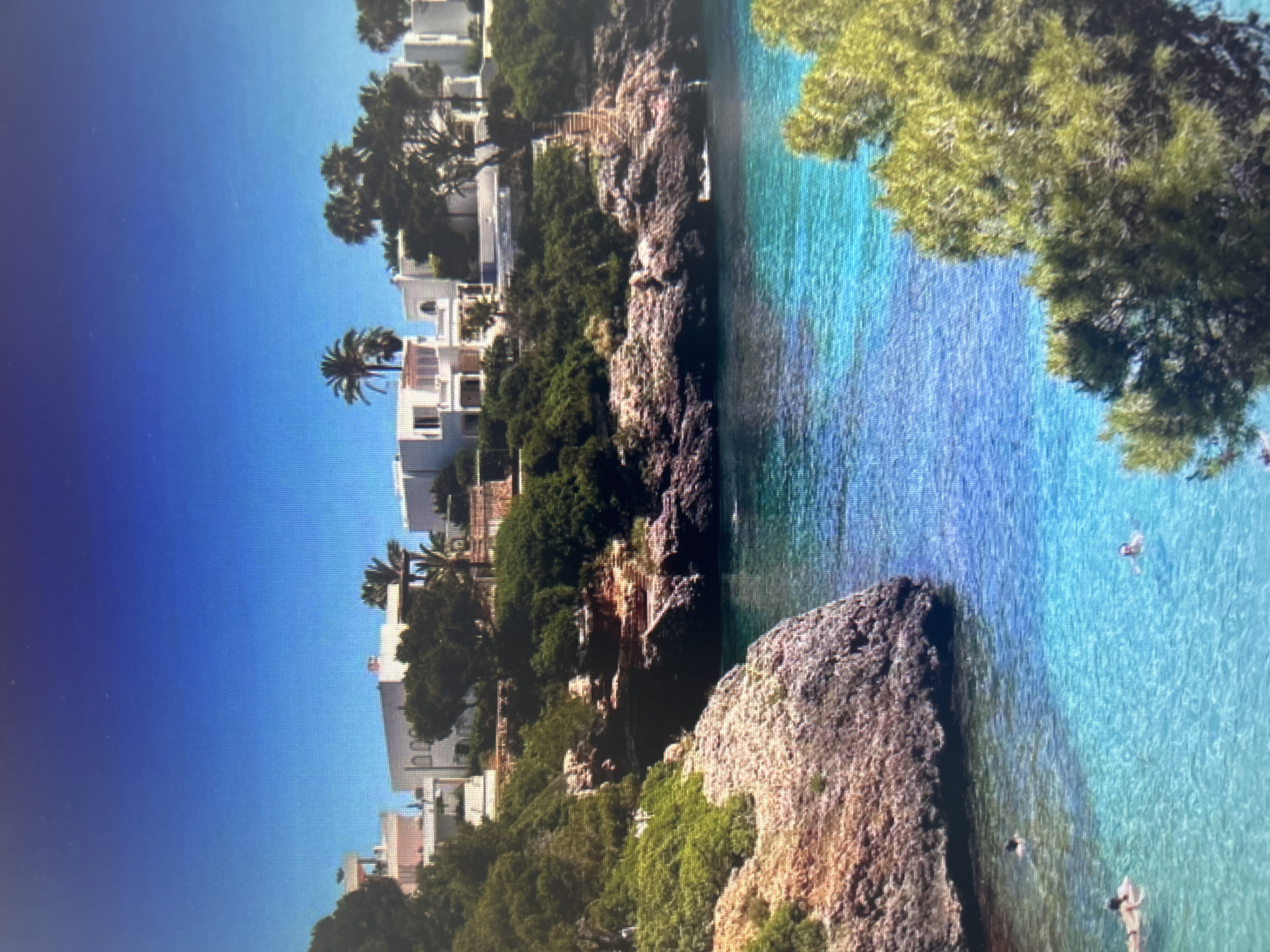 The image depicts a vibrant, sunlit coastal scene, likely a cove or bay. The dominant features are the clear, turquoise-blue ocean water, rocky cliffs, lush green vegetation, and numerous white buildings nestled along the coastline.
In the foreground and middle ground, the pristine water reveals the rocky seabed in places, indicating its clarity. Several small figures are visible in the water, appearing to be swimming or floating, enjoying the calm conditions. Rocky outcroppings and larger boulders extend into the water, with one prominent rock formation in the mid-right.
The shoreline consists of rugged, dark brown and reddish-brown cliffs, heavily covered with various green trees and bushes. Scattered among this greenery, particularly on the left side of the frame, are several white, multi-story buildings, possibly houses or hotels, with some featuring balconies. Palm trees and other evergreen trees are prominent, adding to the tropical or Mediterranean feel.
The sky above is a clear, bright blue, fading slightly to a lighter hue near the horizon, suggesting a sunny day with excellent weather. The overall impression is one of a beautiful, tranquil vacation spot during daytime hours. There is no visible text in the image.