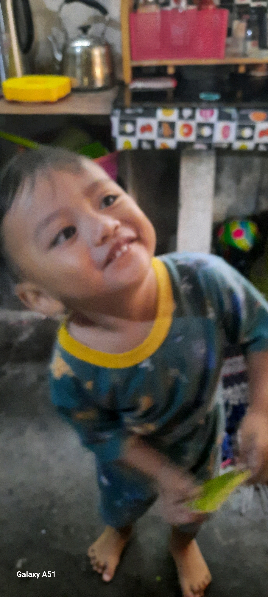 In Jatitengah, Indonesia, a young child, likely a toddler, is the primary subject of this indoor photograph. The child is wearing a teal shirt with a yellow collar and a pattern of yellow motifs, possibly stars or flowers. They are positioned in the lower half of the frame, looking towards the camera with a broad smile, revealing visible teeth. The child's face is the sharpest element in the image, while the rest is out of focus. The background suggests a domestic setting, with shelves holding various items, including a silver kettle and a pink bin. A patterned fabric hangs on the shelf below, decorated with colorful circles and shapes. The floor appears to be a rough, concrete-like surface. The child's bare feet are visible at the bottom of the frame, and they are holding a bright green object in their right hand. The lighting is somewhat dim, creating a slightly hazy atmosphere. The bottom left corner of the image displays "Galaxy A51", indicating the device used for the photograph.