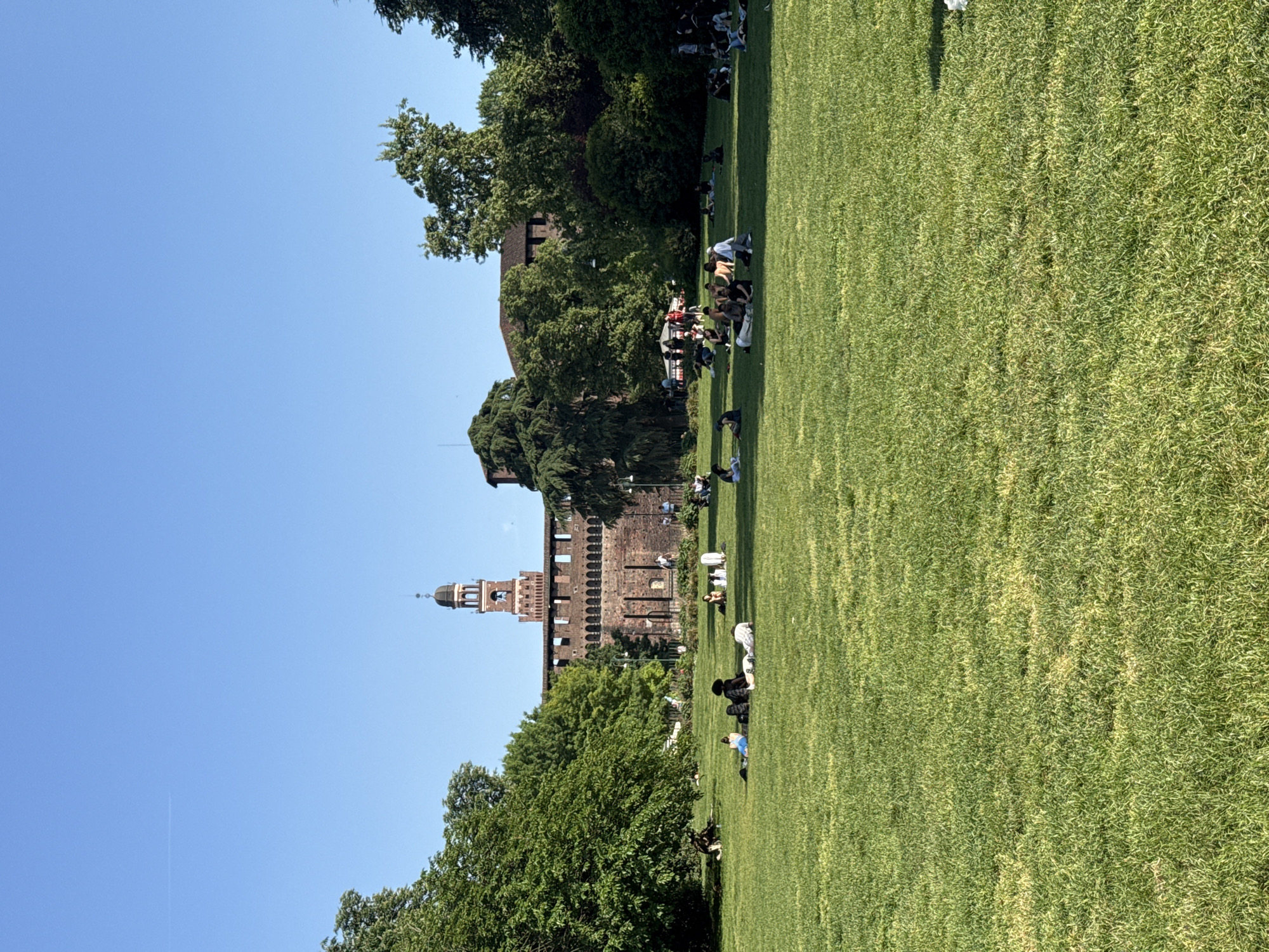 A wide shot captures a sunny day in a large, verdant park, with a historic castle visible on the left side of the frame. The image is oriented vertically with the sky and treetops on the right and the grassy field and castle on the left. For clarity, I will describe it as if correctly oriented horizontally.
The scene depicts a clear, bright blue sky dominating the upper portion of the image, with sparse white contrails suggesting an aircraft has passed. Lush green trees frame the top and bottom left of the photo, partially obscuring the view of the castle.
In the mid-ground, the prominent feature is a large, light brown brick castle or fortress, identified as the Sforza Castle in Milan, Italy. Its distinctive architecture includes battlements along the walls and a tall, multi-tiered clock tower (Torre del Filarete) topped with a dome, which rises against the clear sky.
The foreground and much of the right side of the image are covered by an expansive, well-maintained green lawn. Numerous people are scattered across the grass, enjoying the sunny weather. They are mostly seen sitting or lying down, individually or in small groups, suggesting a leisurely and relaxed atmosphere. Some individuals are visible near the castle walls, while others are further out in the open parkland.
The overall impression is one of a peaceful, pleasant afternoon in a public park, with visitors unwinding near a significant historical landmark. No specific text is visible in the image.