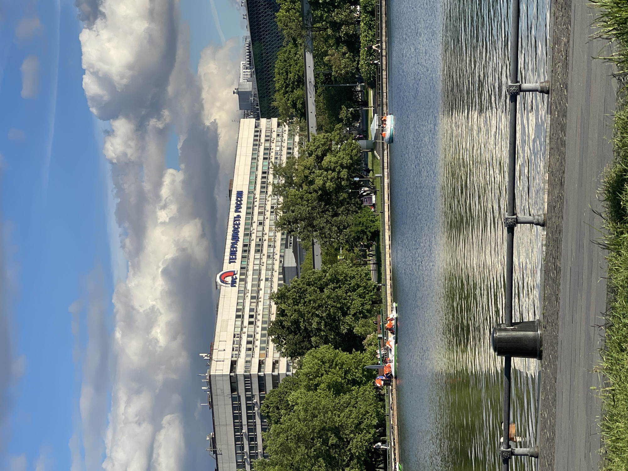 The image shows a vertical view of a scene featuring a body of water, a building, and a cloudy sky. The body of water occupies a significant portion of the right side of the image, with ripples visible on its surface. Along the edge of the water is a paved walkway with metal posts.
On the left side of the image, a tall, white building is prominent. The words "ТЕЛЕРАДИОКОМПАНИЯ РОССИЯ" are written vertically in blue on the building's facade, with a red and white logo above the text. Behind the building, a modern structure with a glass facade can be seen. Several trees with green foliage fill the space between the building and the walkway.
The sky is filled with a combination of blue and white clouds, with some clouds appearing darker and more ominous. The overall lighting suggests a daytime scene with partly cloudy conditions. The image appears to be taken in Moscow, Russia.