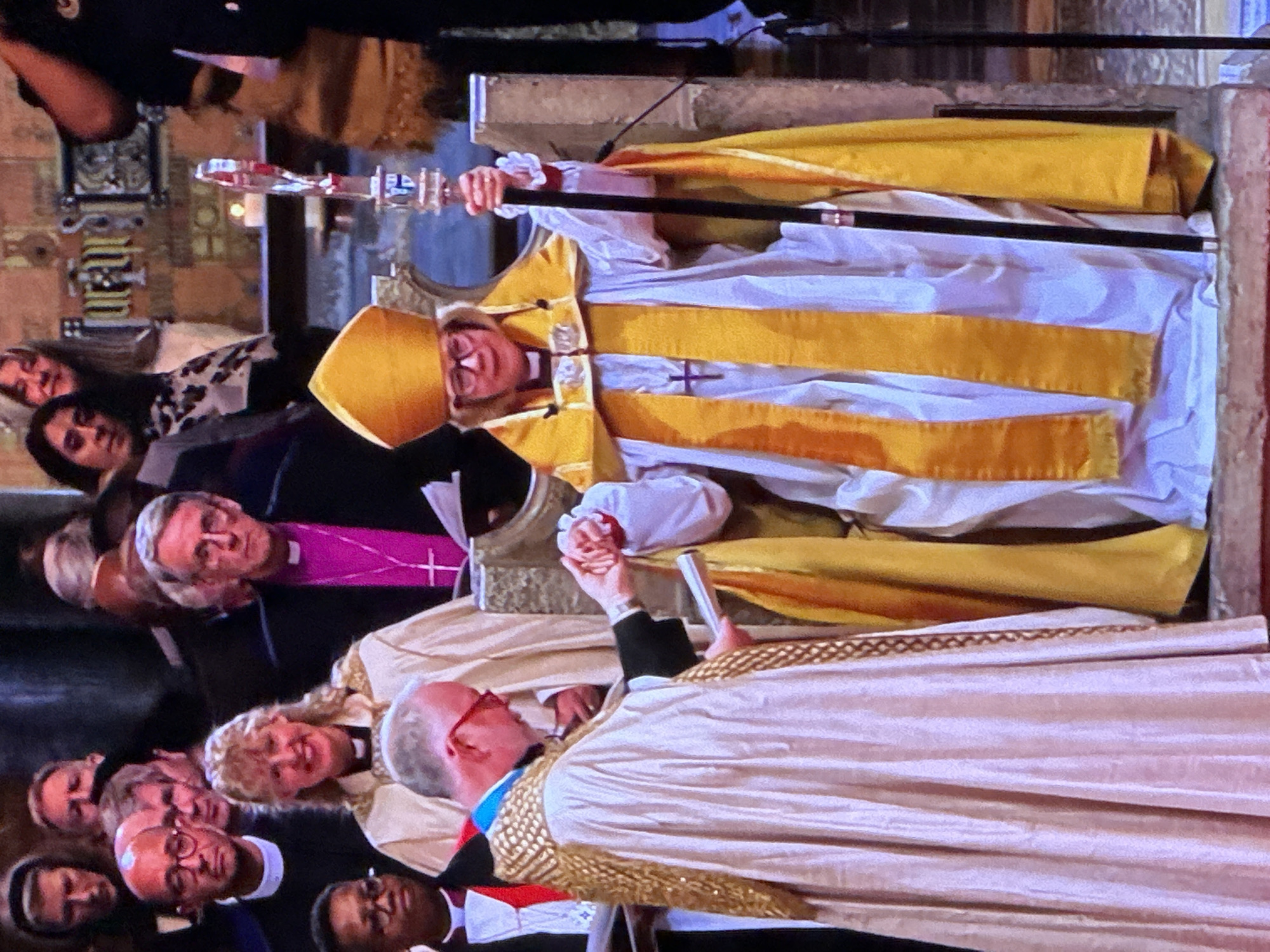 The image depicts a formal religious ceremony taking place inside St Paul's Cathedral, London, United Kingdom, characterized by its stone architecture visible in the background. In the foreground, an older woman, identified as a bishop, is seated on a ceremonial stone chair. She wears a white alb, a bright yellow stole, a yellow cope, and a golden mitre, holding a black crozier with a clear decorative top in her right hand. Her right hand is grasped by an older man standing before her, who wears a light-colored alb and a gold-patterned cope over a dark clerical outfit. He holds a scroll or book in his left hand. Both central figures wear glasses. Behind them, numerous individuals observe the ceremony; some wear clerical attire, including black cassocks and clerical collars, while others are in formal civilian clothing. The background includes decorative stone carvings and a dimly lit area with a single lit candle visible on a ledge. The scene captures a solemn interaction between the two central figures, indicative of a consecration or installation rite.