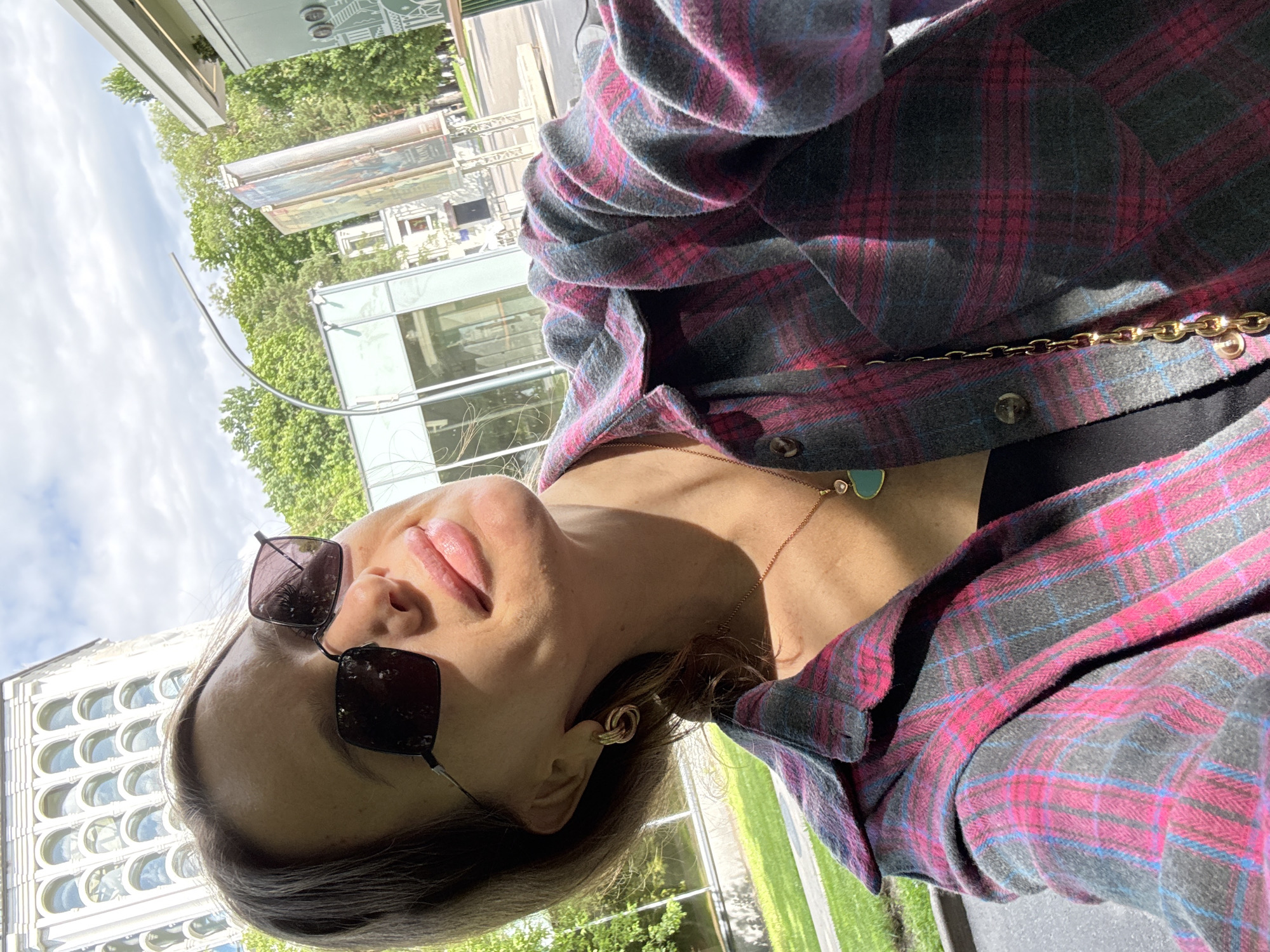 A woman is taking a selfie outdoors in Moscow, Russia, under a partly cloudy sky. She is wearing a plaid shirt, sunglasses, and a necklace with a heart-shaped pendant. In the background, there is a large, ornate building with arched windows, as well as trees and other urban structures. The sun is shining, casting shadows on her face and shirt.