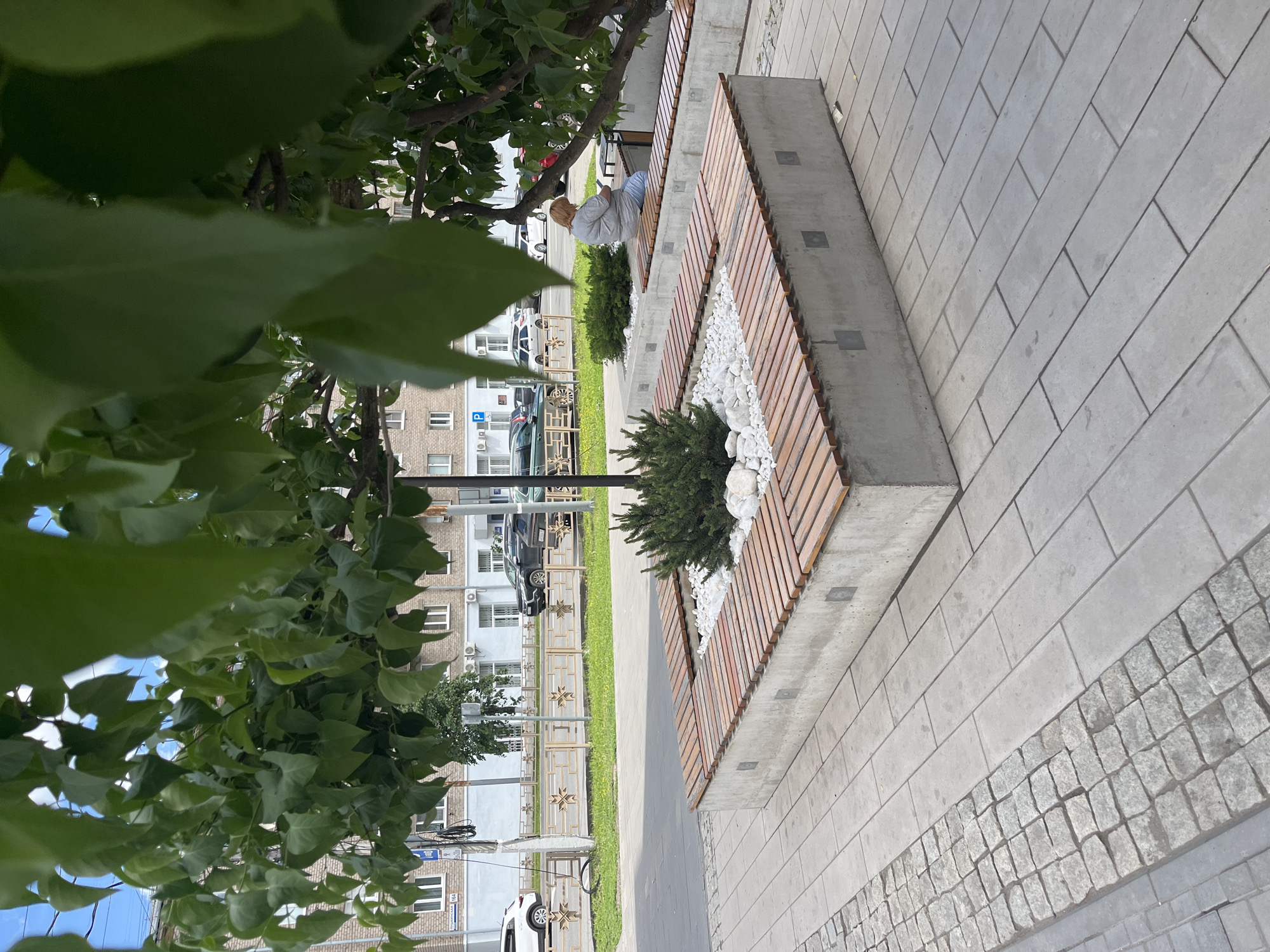 A street scene in Izhevsk, Russia, shows a concrete and wood bench arrangement with a central decorative rock and shrub planter. A gray-haired person in a gray shirt sits at the far end of the bench. Paved walkways surround the bench. Trees are visible at the foreground left, with a building visible in the background across a narrow strip of grass. Several parked cars, a street lamp, and a metal fence run along the street in the background.