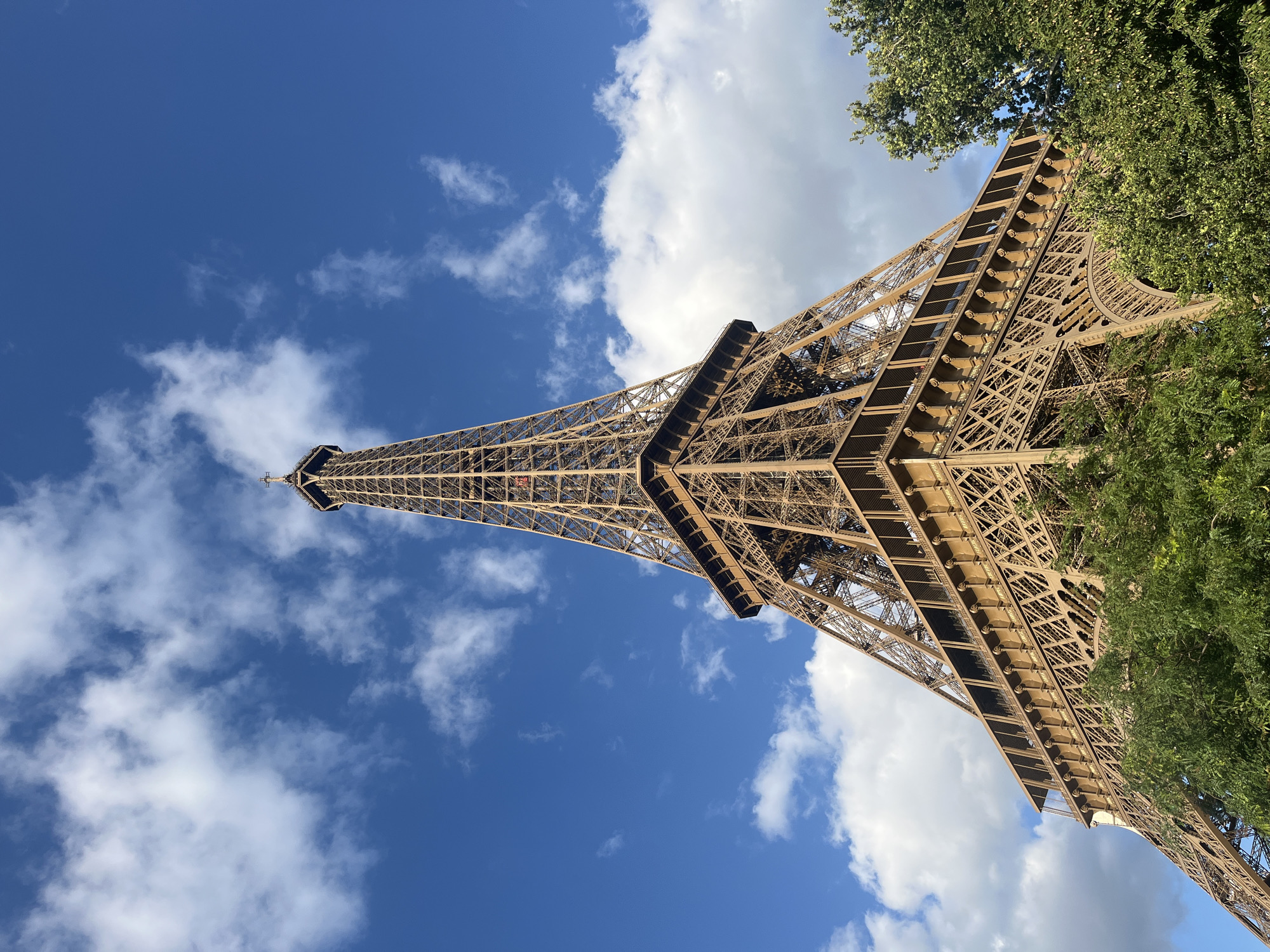 The image is a low-angle, close-up shot of the Eiffel Tower in Paris, France, against a vibrant blue sky dotted with white clouds. The iconic iron lattice structure of the tower dominates the frame, its intricate metalwork visible in detail. The lower portion of the tower is partially obscured by lush green trees, suggesting it is a sunny day. The perspective emphasizes the immense height and scale of the landmark. There are no people or active events depicted in the scene; it is a static, architectural study of the monument.