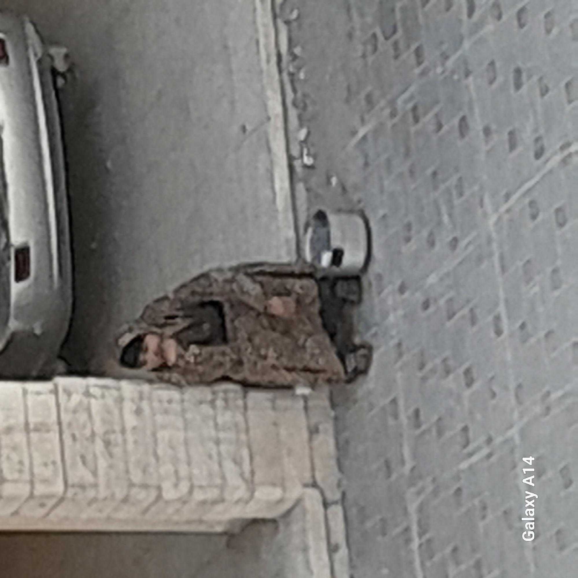 The blurry, low-resolution image, apparently taken with a "Galaxy A14" phone (text visible along the right edge), captures a somber scene in what appears to be an urban or semi-urban outdoor setting.
The main subject is a person lying down, seemingly asleep or resting, on a makeshift bed of what looks like blankets or discarded materials. Their head and upper body are partially visible, with a light-colored face and hands suggesting a fair complexion. They are positioned on a raised concrete or stone ledge, which forms part of a building's structure, possibly a balcony base or a large windowsill.
To the left, a portion of a light-colored vehicle, likely a car, is visible. Its rear fender and part of a taillight are distinguishable. Above the person's head, on the ground or a surface next to them, rests a small, light-colored metal can or container. The wall of a building, characterized by a repeating, textured, possibly brick-like or stucco pattern, occupies the right side of the frame. The lighting is diffused and flat, suggesting either an overcast day or an area with limited direct sunlight, such as an alleyway.
The overall scene conveys a sense of vulnerability and hardship, possibly depicting someone experiencing homelessness seeking rest in an exposed public space. The image quality is poor, with significant pixelation and blur, making fine details difficult to discern, but the general elements of the scene are clear.
