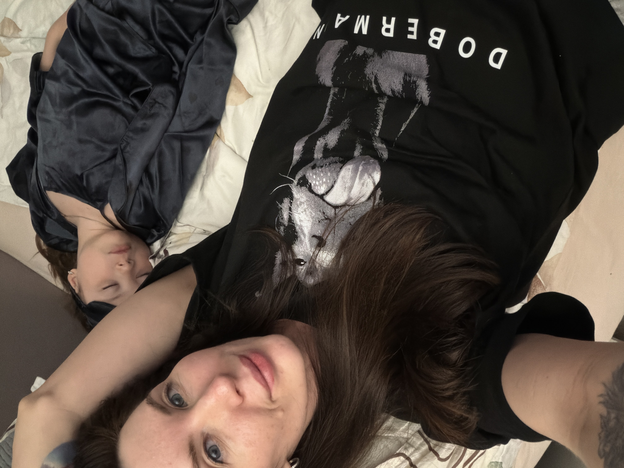 This image, taken indoors in Perm, Russia, depicts two individuals resting on a bed. In the foreground, a person is taking a selfie, looking directly at the camera with blue eyes and a calm expression, possibly a slight smile. They have long, dark brown hair and are wearing a black t-shirt that features a graphic print of a Doberman dog's head. Above the dog's head, the word "DOBERMAN" is visible in white, appearing upside down from the viewer's perspective. A tattoo is discernible on their left arm.
Lying beside this individual, further back and to the left, is another person who appears to be asleep or deeply relaxed. Their eyes are closed, and their face is serene. They are dressed in a dark, possibly satin, nightgown or robe. The bed is covered with light-colored bedding, featuring a subtle pattern. The overall scene conveys a sense of peaceful relaxation, likely during leisure time.